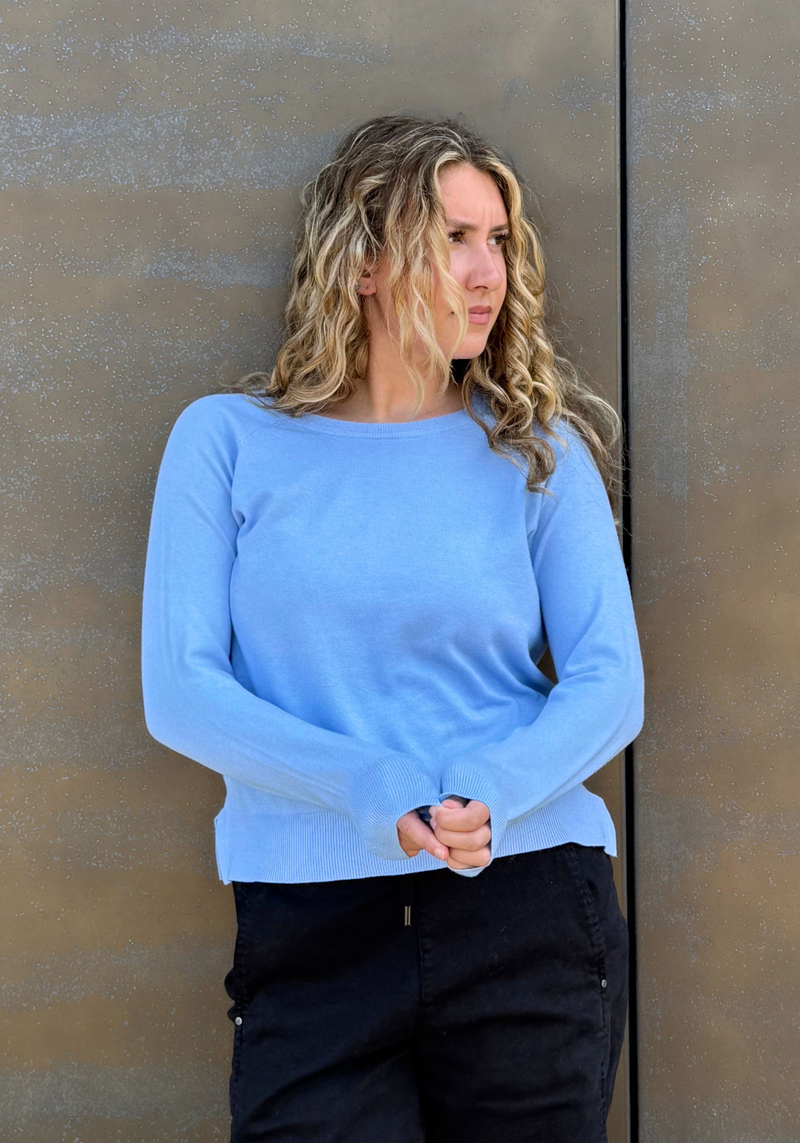 A woman wearing a light blue long-sleeve knit top and black pants, standing against a textured brown wall.