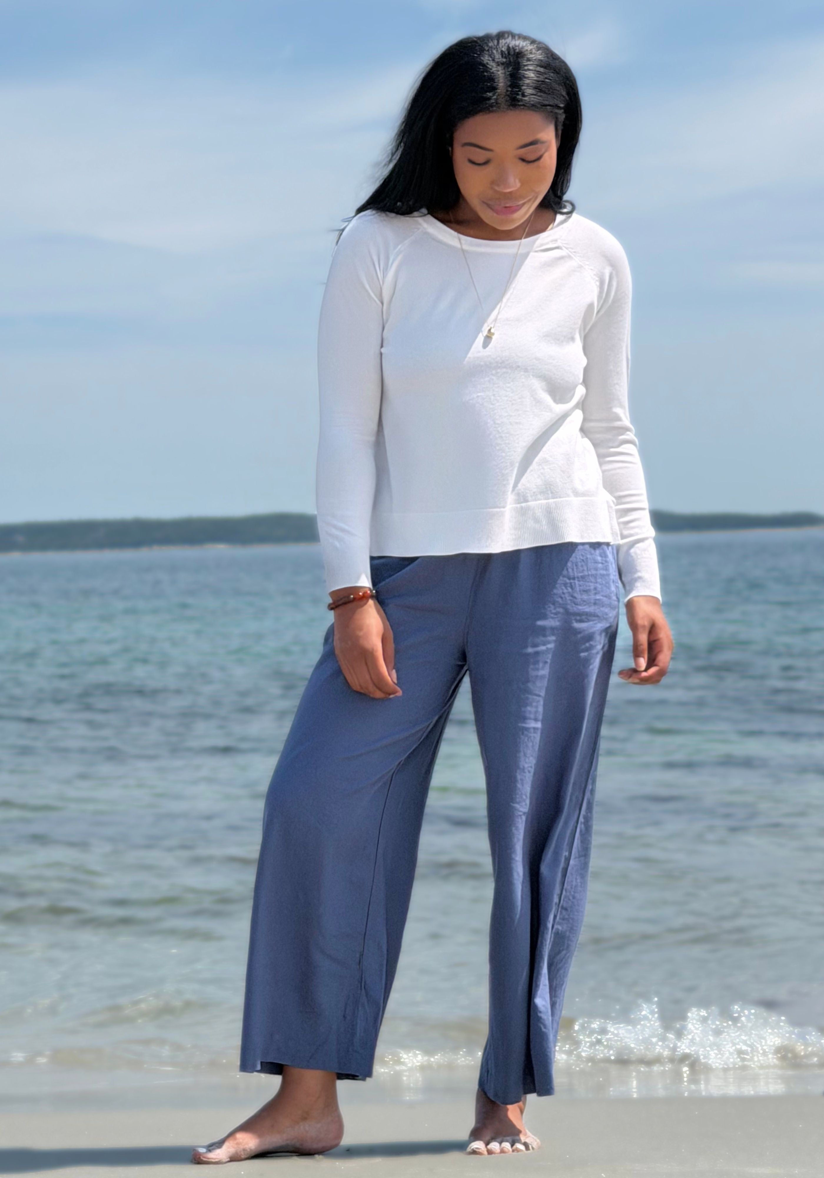 A woman wearing a white long-sleeve top and slate blue wide-leg pants, with the ocean and sky in the background.