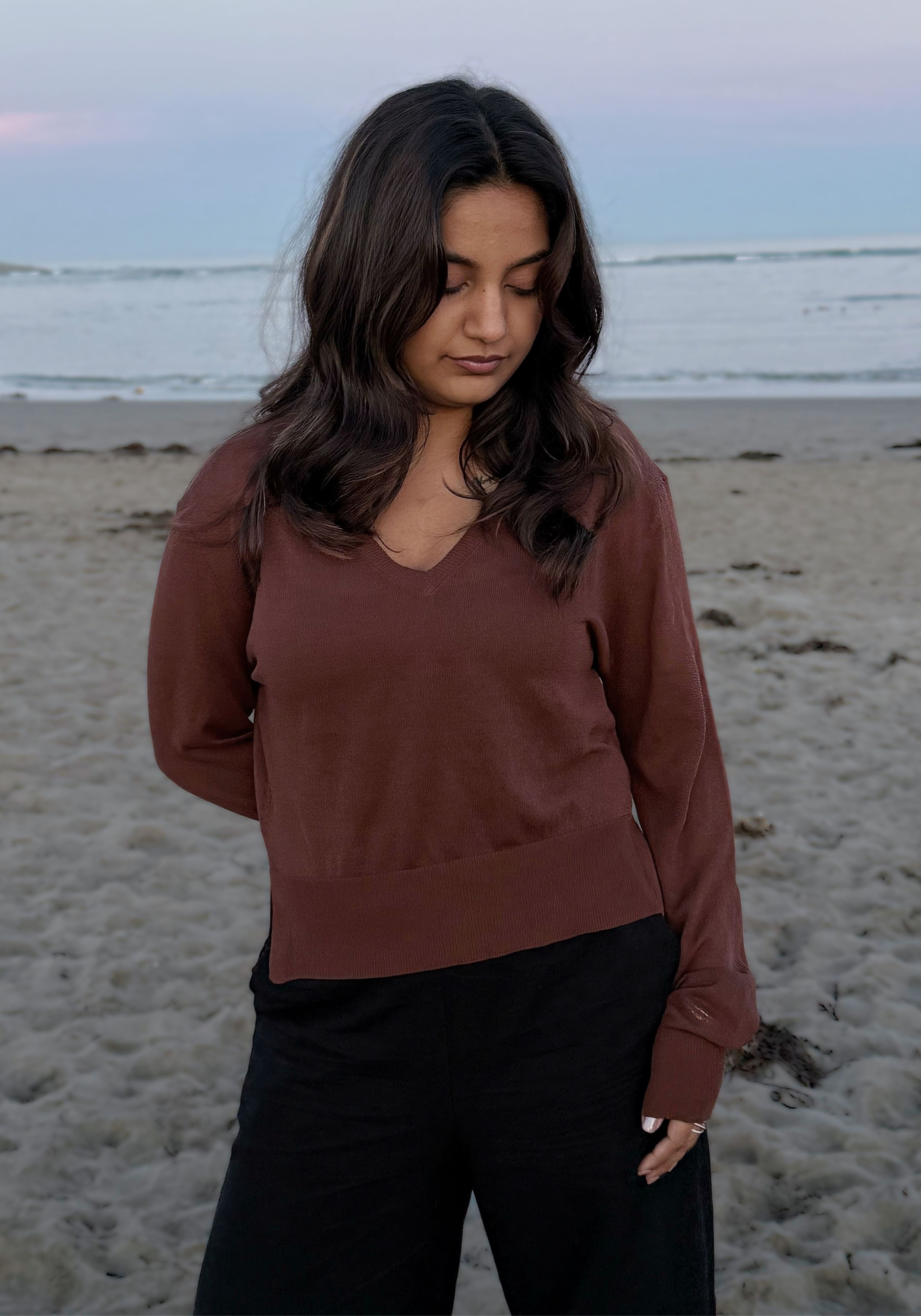 A woman standing on a sandy beach wearing a rust-brown long-sleeve V-neck top and black pants.