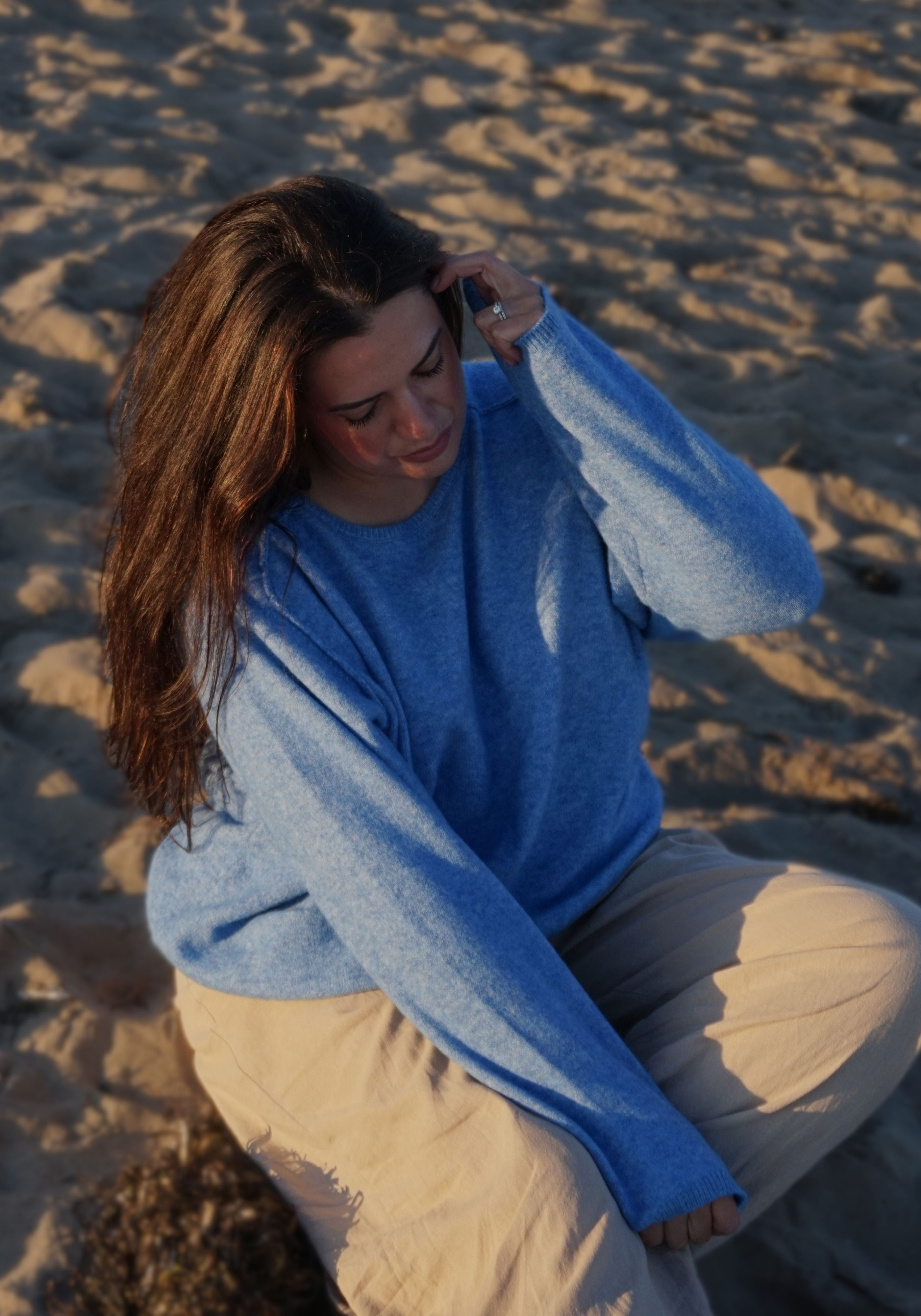 A woman with long brown hair sits on a sandy beach wearing a light blue knit sweater and beige pants, looking down while holding a phone to her head.