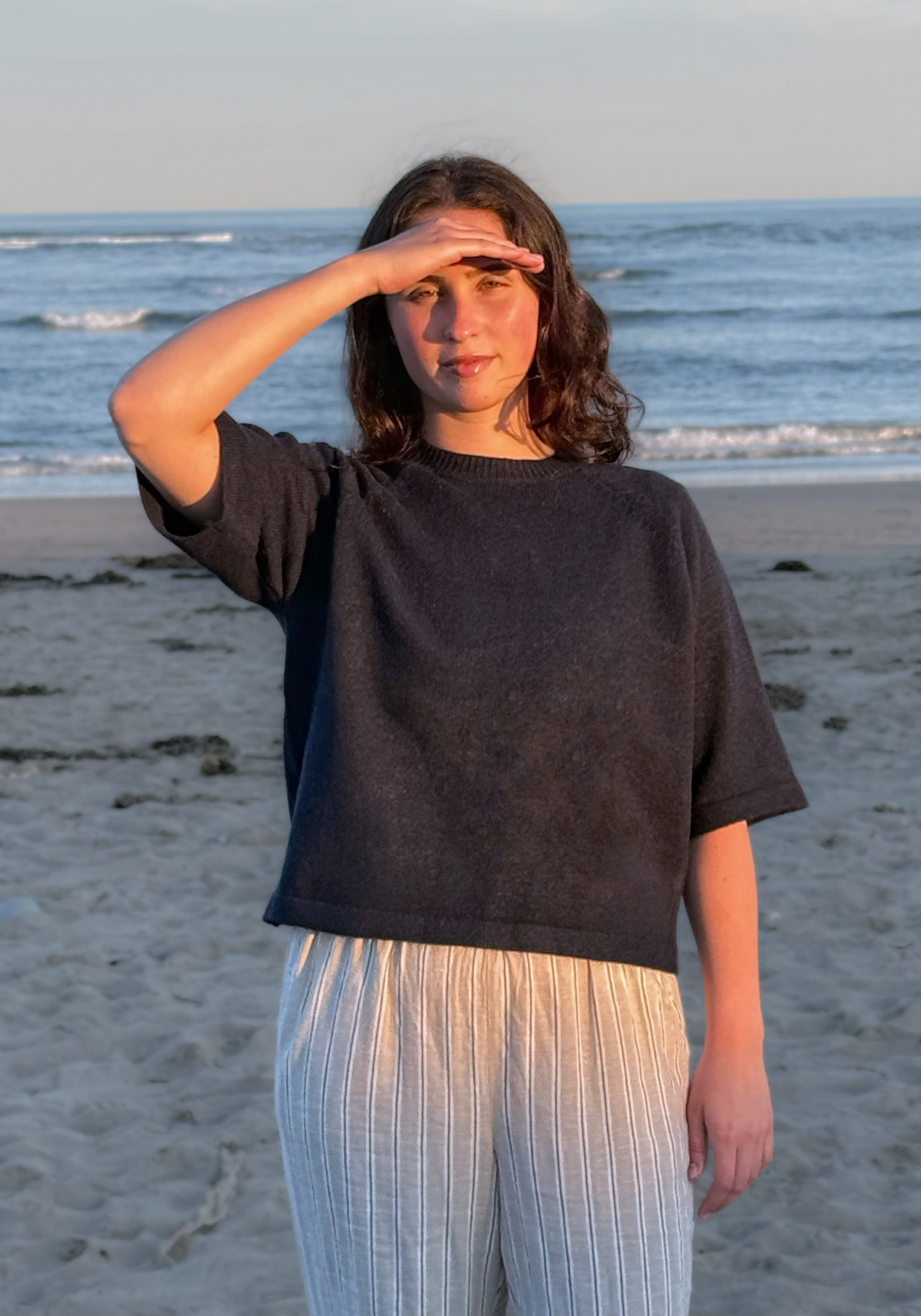 A woman standing on a sandy beach with the ocean in the background, wearing a dark charcoal gray oversized short-sleeve knit top and light beige vertically striped pants.