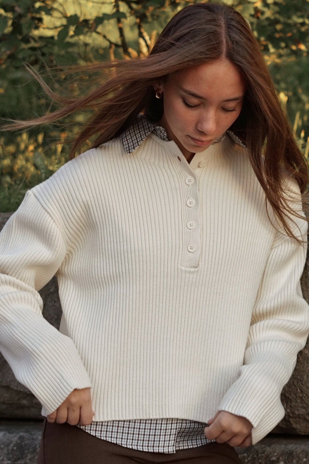 A young woman wearing a cream ribbed knit button-up henley sweater with a plaid collar detail, standing outdoors against a stone wall with green foliage in the background.