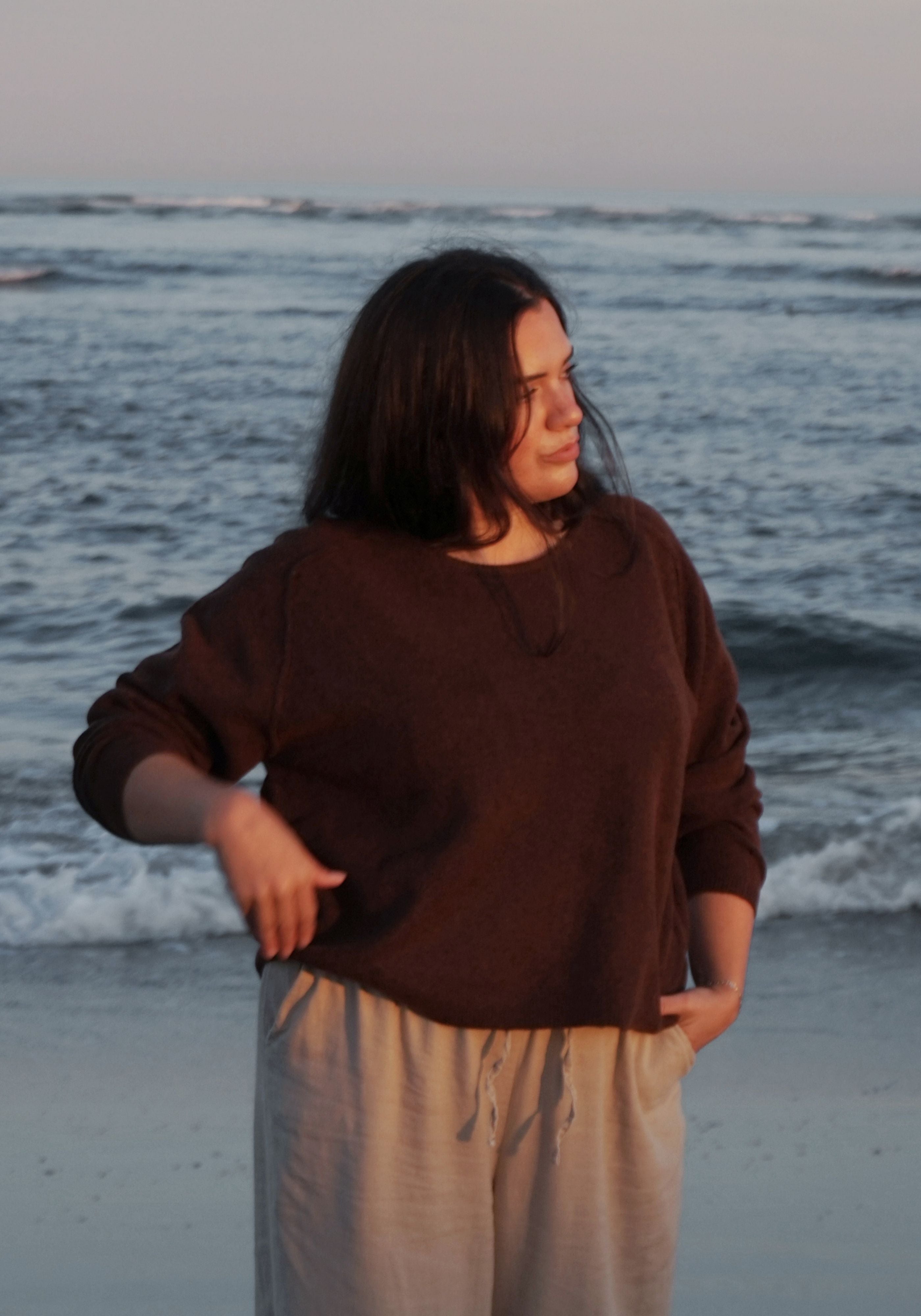 A woman standing on a beach wearing a dark brown long-sleeve knit top and light beige drawstring pants, with ocean waves in the background.