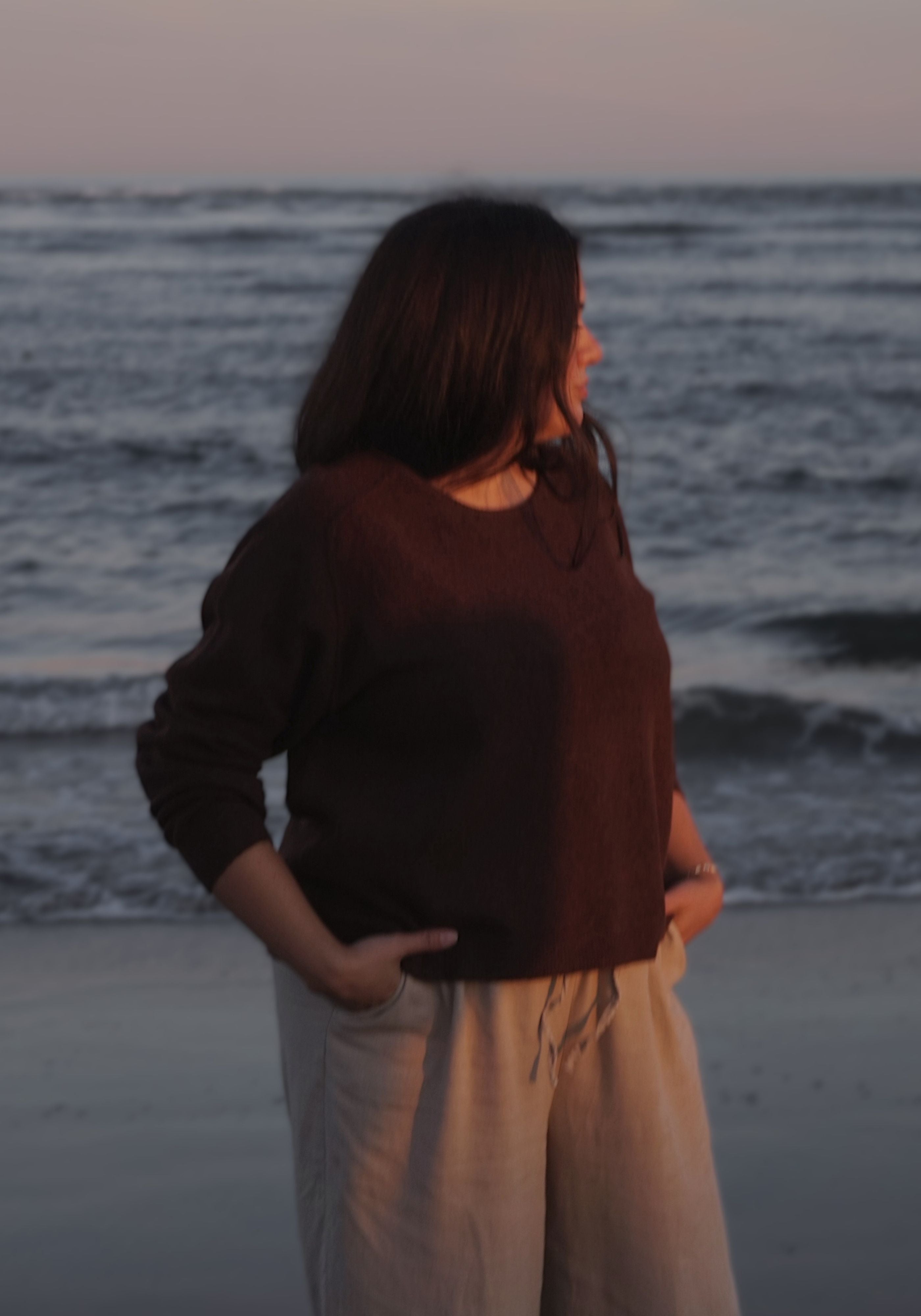A woman standing on a beach at sunset, shown from the back and side, wearing a dark brown long-sleeve top and light beige drawstring pants.