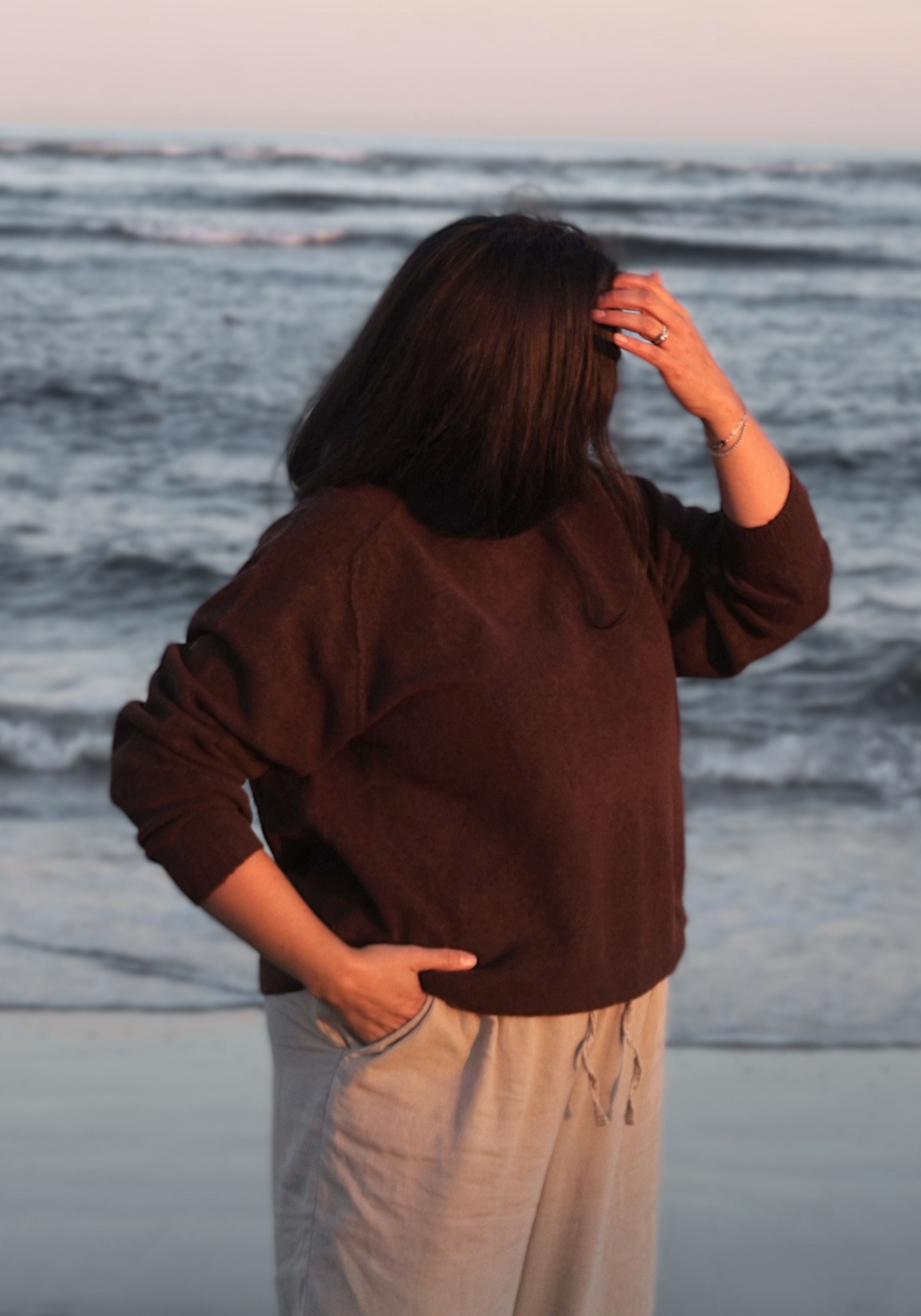 A woman standing on a beach with her back to the camera, wearing a dark brown long-sleeve knit top and light beige drawstring pants, with ocean waves in the background.