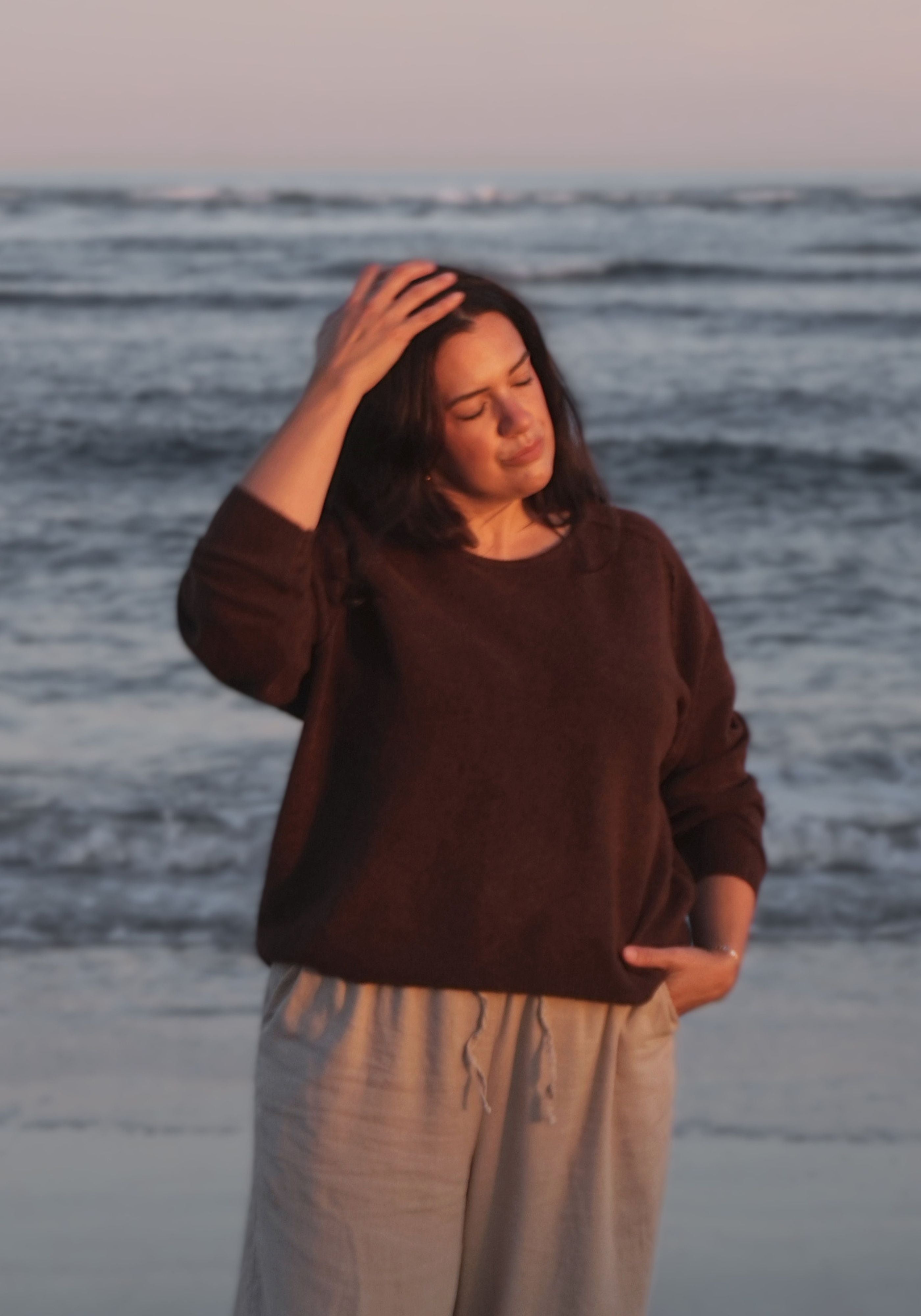 A woman standing on a beach at sunset, wearing a dark brown long-sleeve knit sweater and light beige drawstring pants, with her hand on her head.