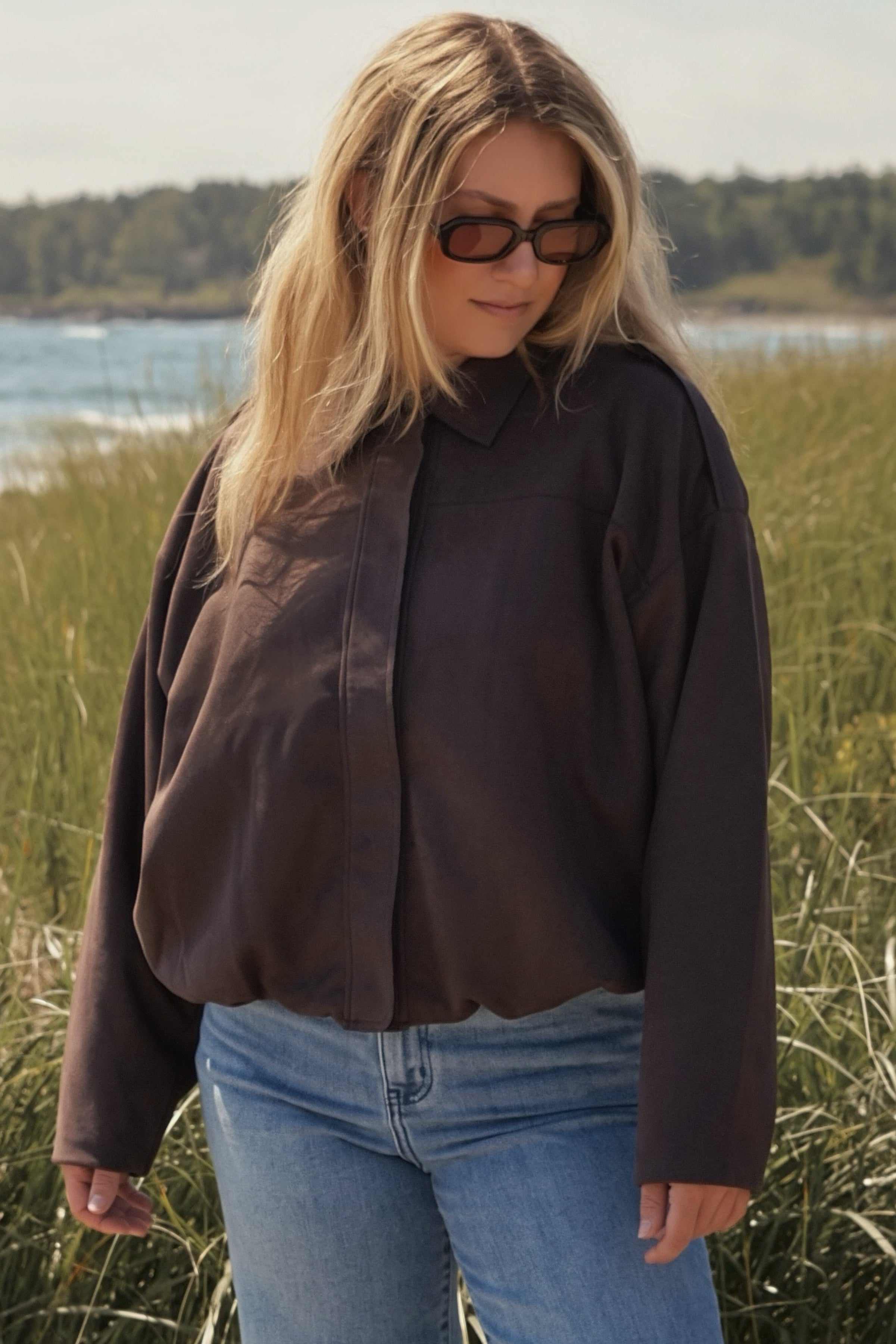 A woman wearing a dark brown button-up shirt and blue jeans stands outdoors in a grassy field near the water, wearing dark sunglasses.
