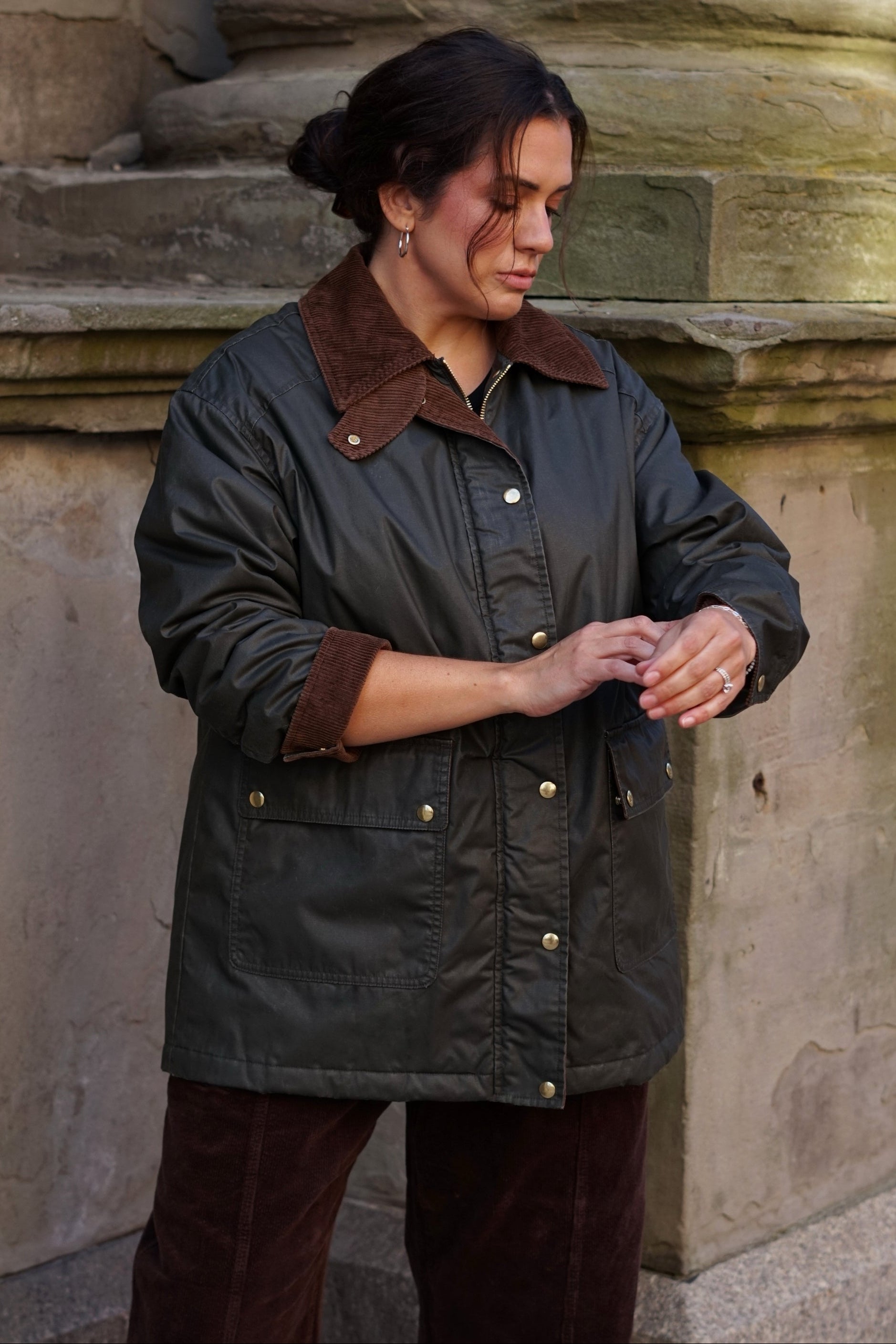 A woman wearing a dark olive green waxed cotton jacket with a brown corduroy collar and cuffs, standing outdoors against a stone wall.