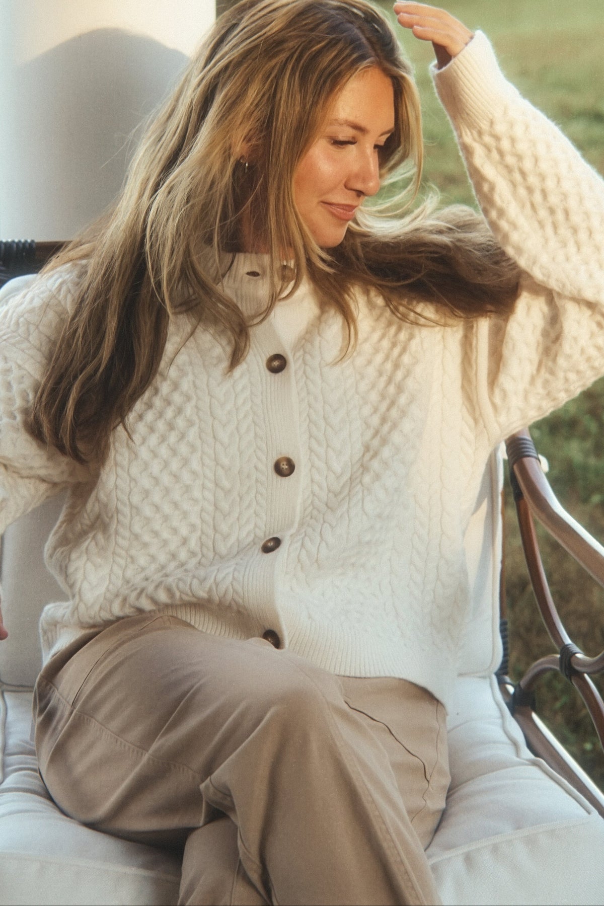 A woman sitting outdoors on a white chair, wearing a cream cable-knit button-front cardigan and beige pants.