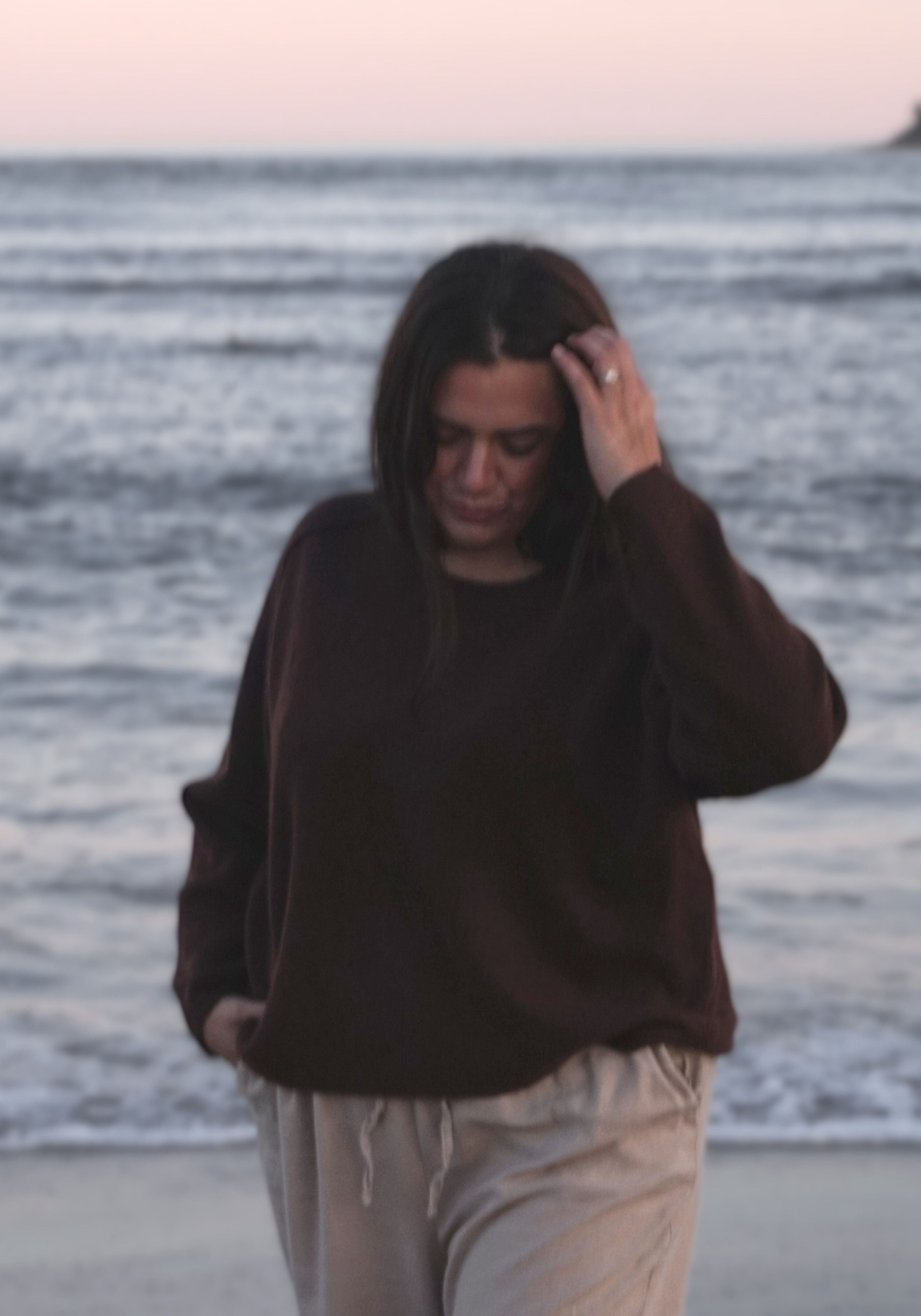 A woman walking on a beach at dusk, wearing a dark brown long-sleeve knit sweater and light beige sweatpants.