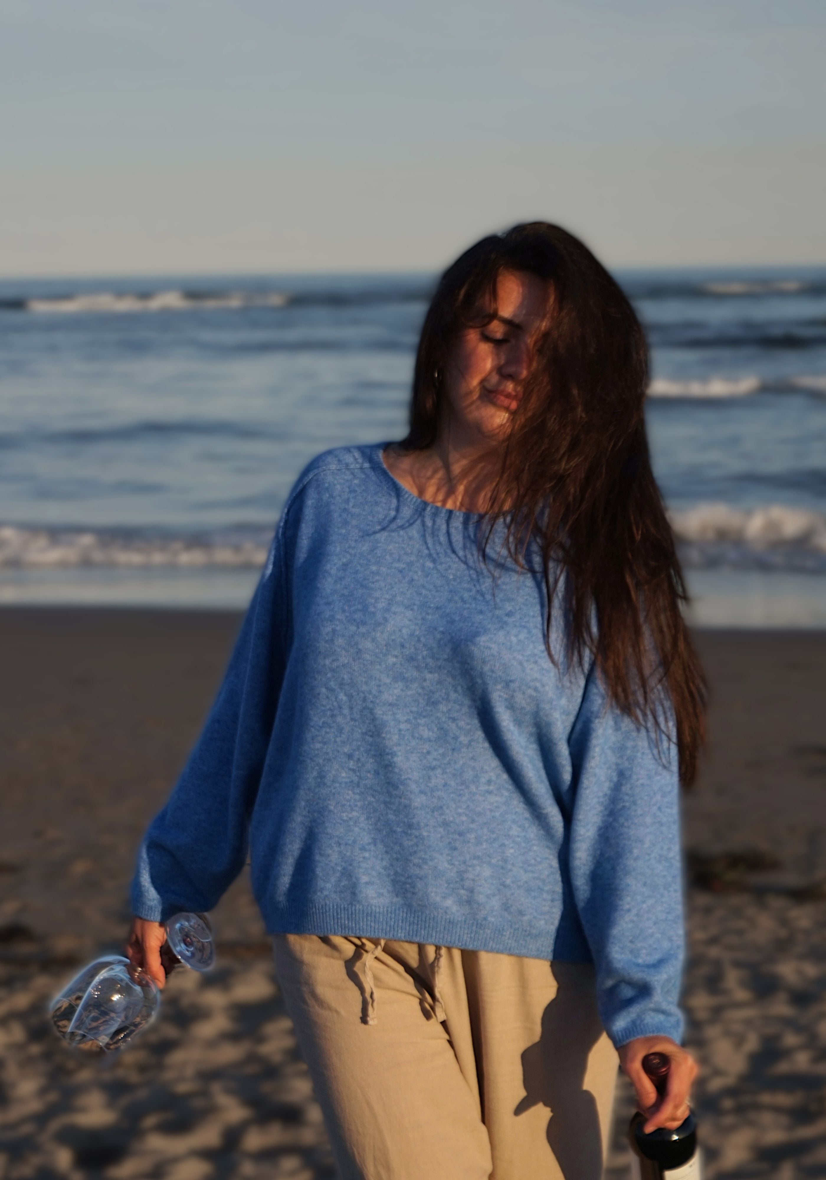 A woman with long dark hair stands on a sandy beach wearing a heathered light blue knit sweater and beige pants, holding wine bottle.