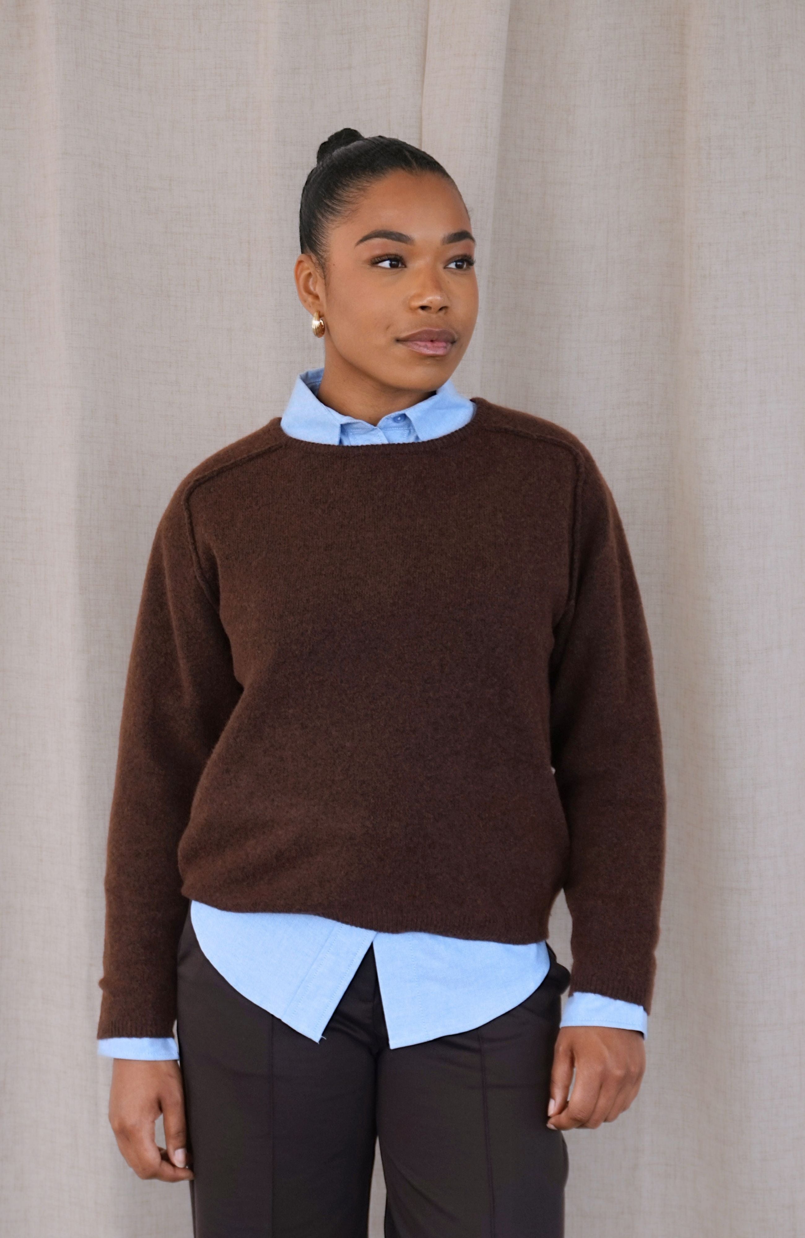 A woman wearing a brown crewneck sweater layered over a light blue collared shirt and dark trousers, standing against a neutral beige curtain backdrop.