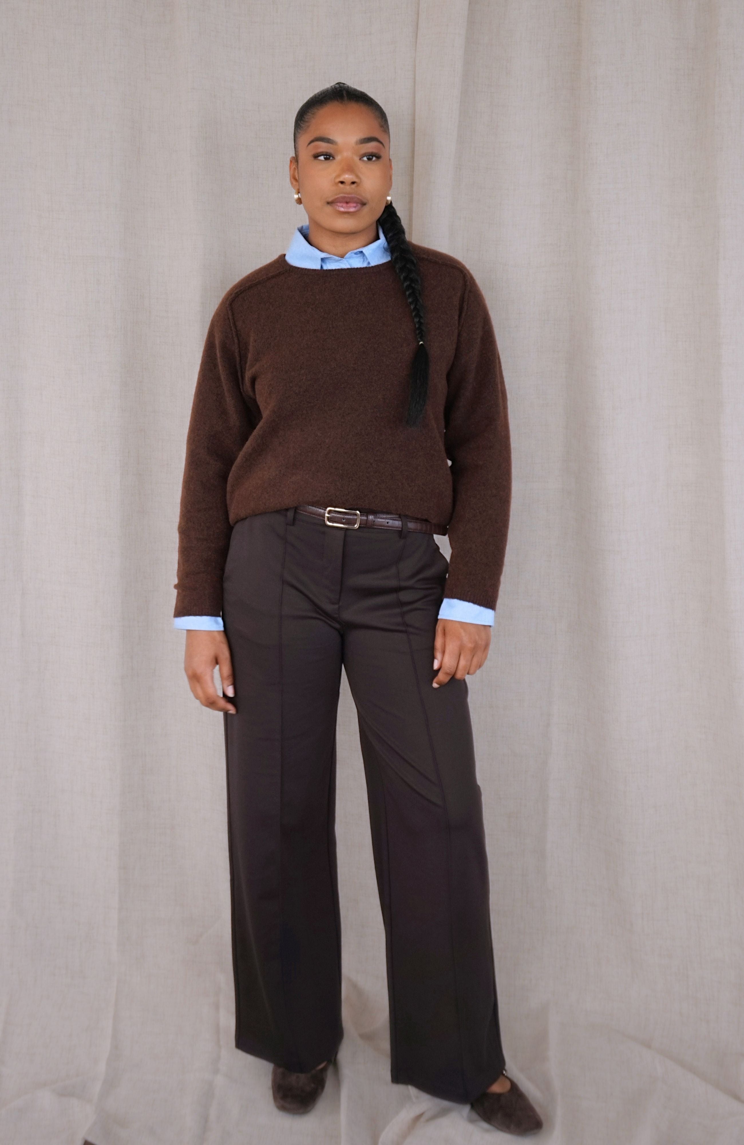A woman stands against a light beige fabric backdrop wearing a brown knit sweater over a light blue collared shirt, dark wide-leg trousers, a dark belt, and dark brown shoes.