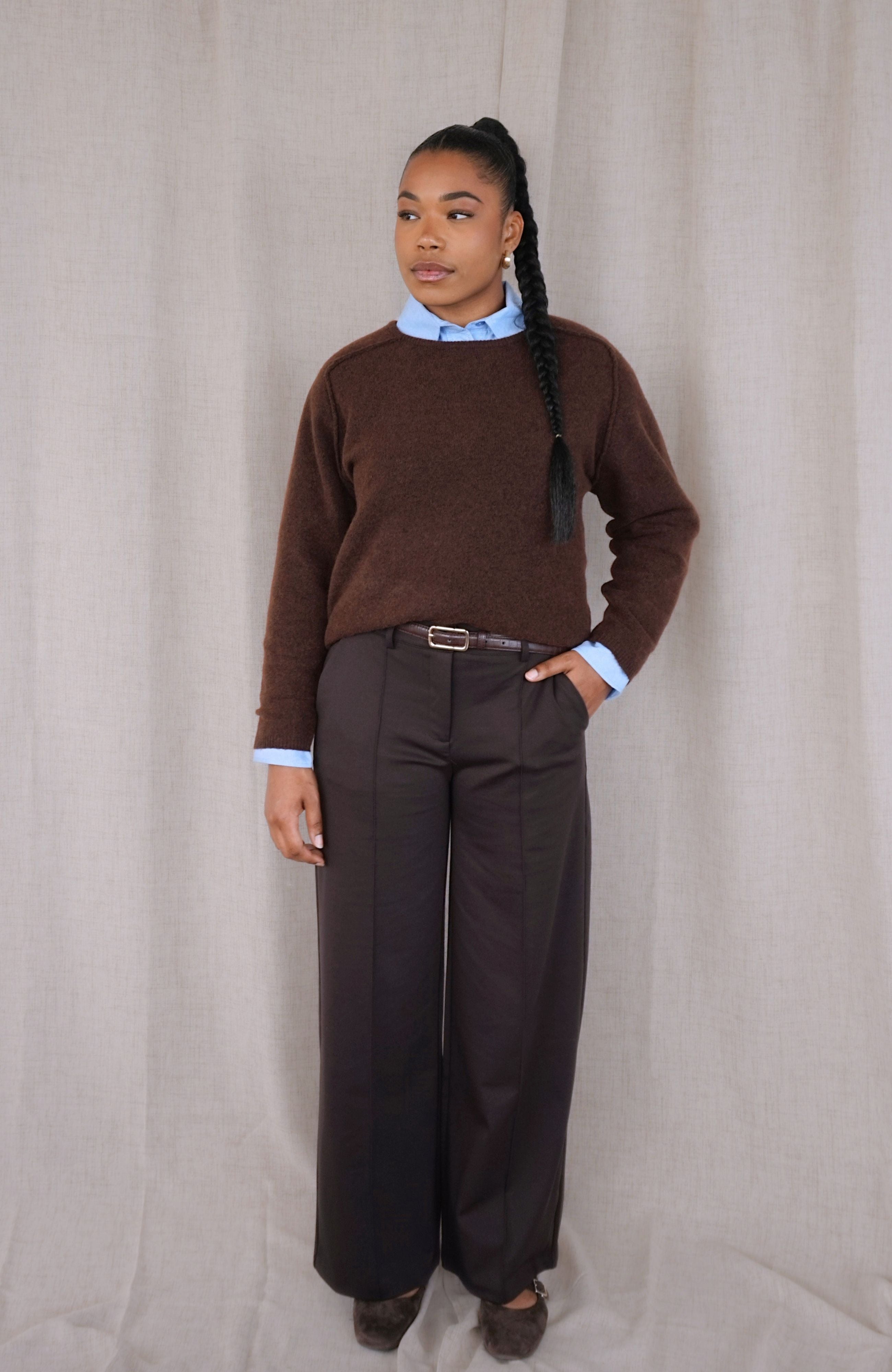 A model stands against a neutral beige fabric backdrop wearing a brown knit sweater layered over a light blue collared shirt, paired with dark charcoal wide-leg trousers and dark shoes.
