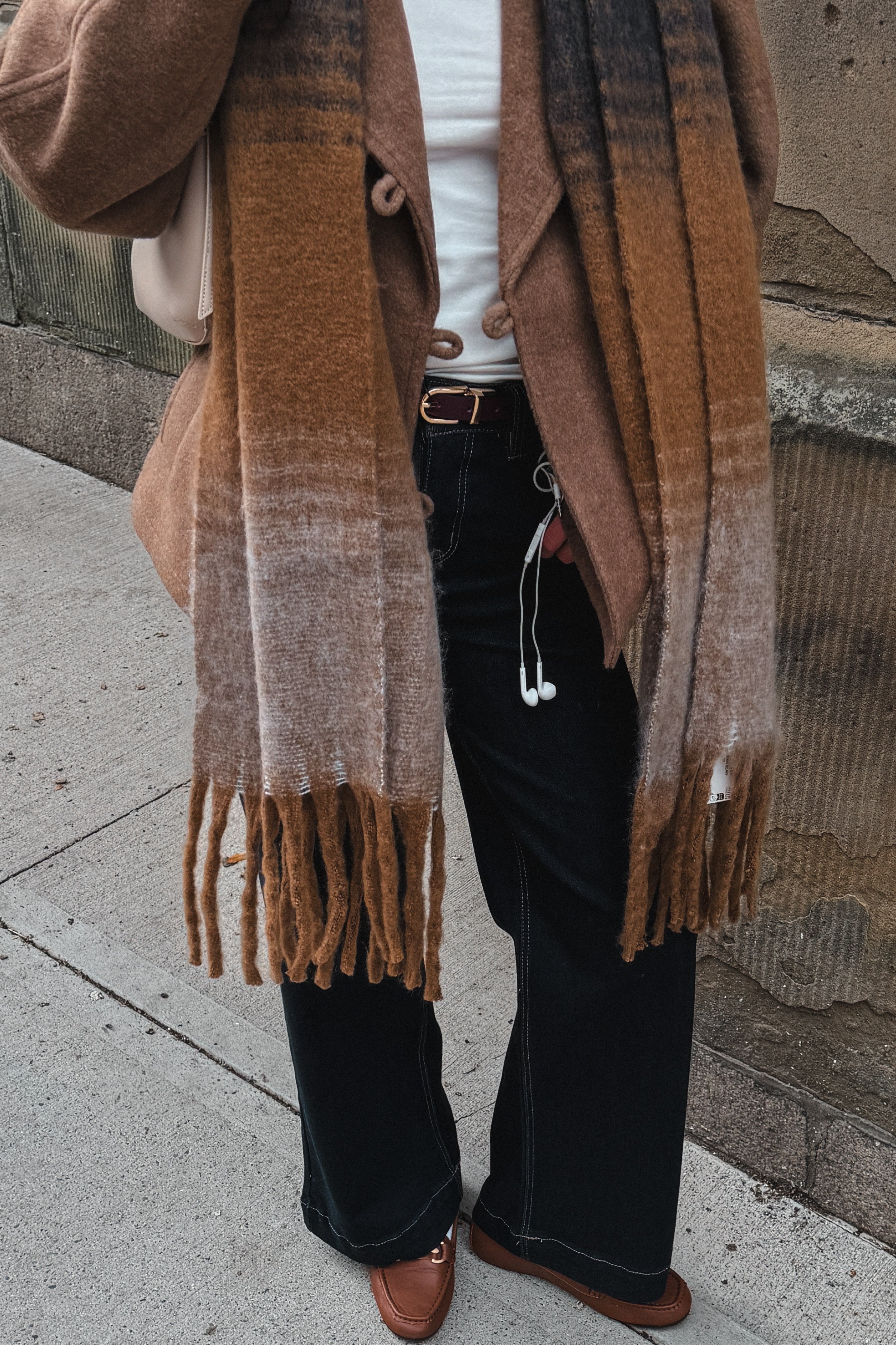 A person standing outdoors on a sidewalk wearing a brown and beige fringed scarf, a tan cardigan, a white top, black wide-leg pants, and brown shoes.