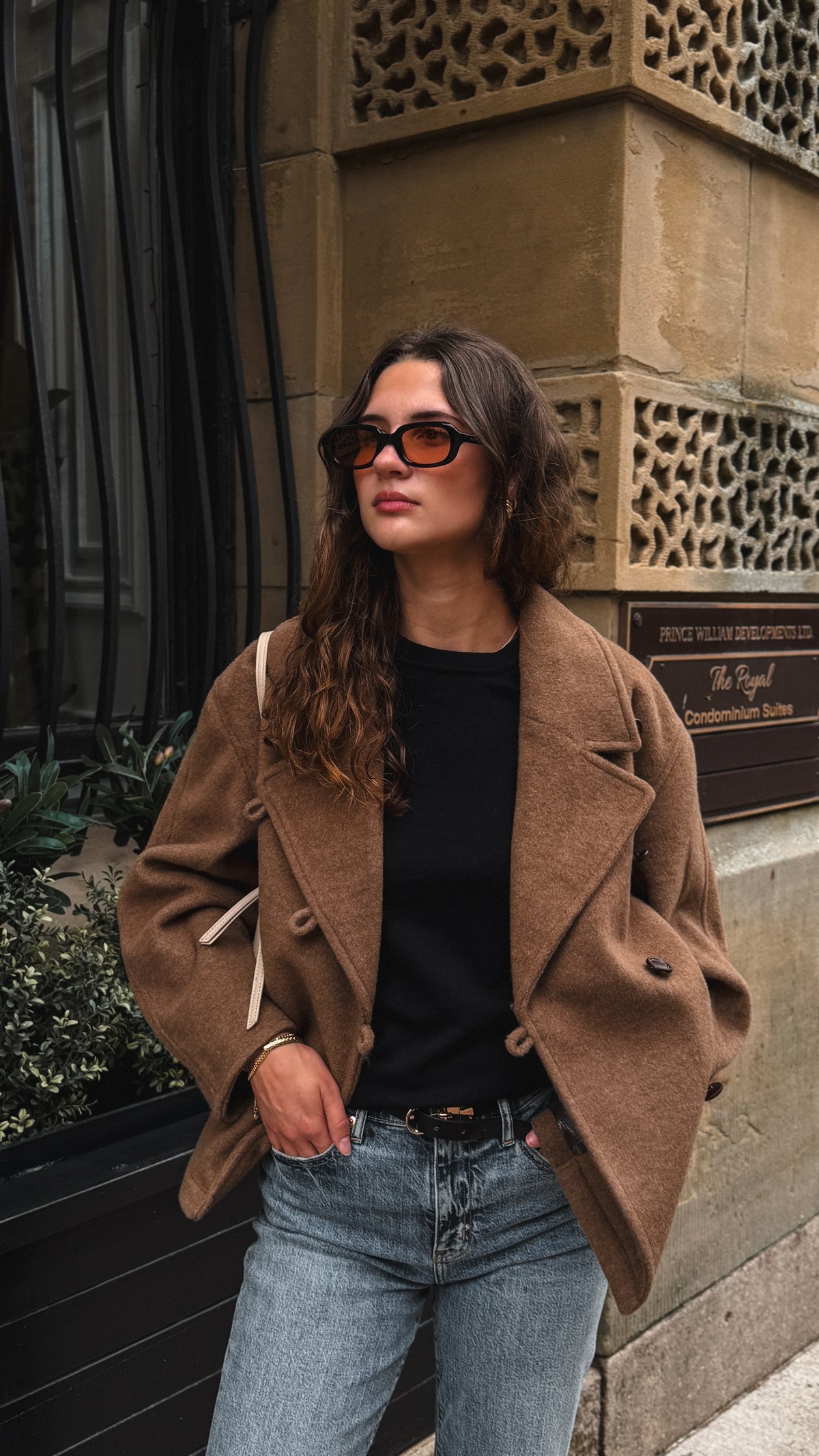 A woman posing outdoors in a brown double-breasted blazer, black top, light-wash blue jeans, and sunglasses, carrying a beige shoulder bag.