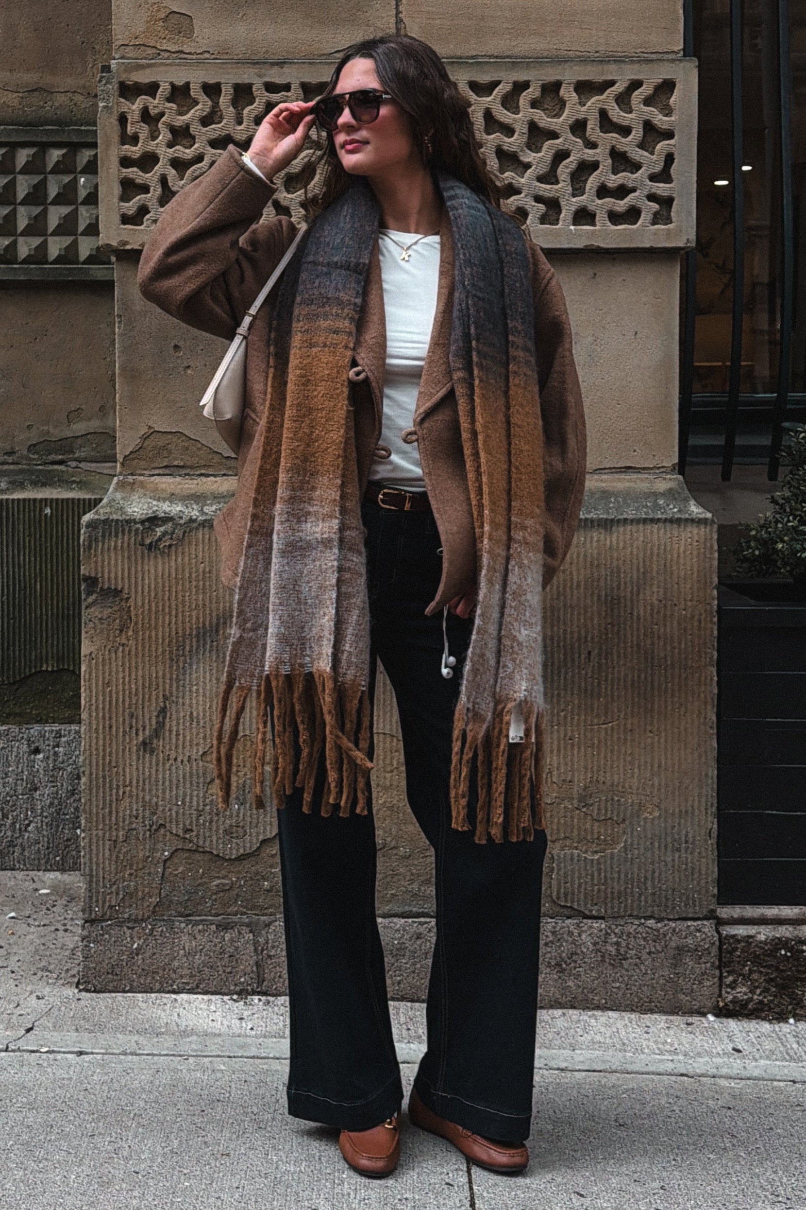 A woman stands on a city sidewalk in front of a stone building, wearing a brown and beige gradient fringed scarf, a brown jacket, a white top, black wide-leg pants, and brown shoes.