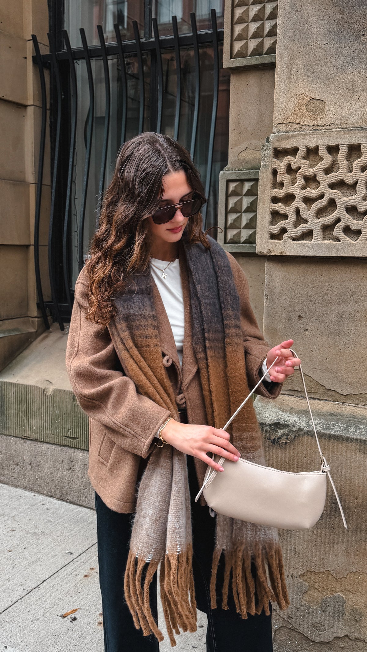 A woman wearing a brown and beige plaid fringed scarf, a brown cardigan, and sunglasses, holding a light beige shoulder bag while standing outdoors against a stone building.