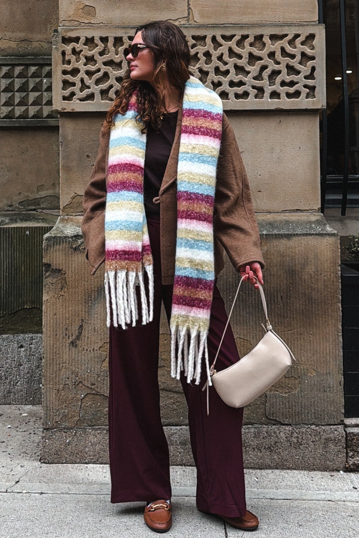 A woman stands on a city sidewalk in front of a stone building, wearing a wide brown coat, dark burgundy wide-leg pants, and a thick multicolored striped knit scarf with white fringe. She carries a light beige shoulder bag.