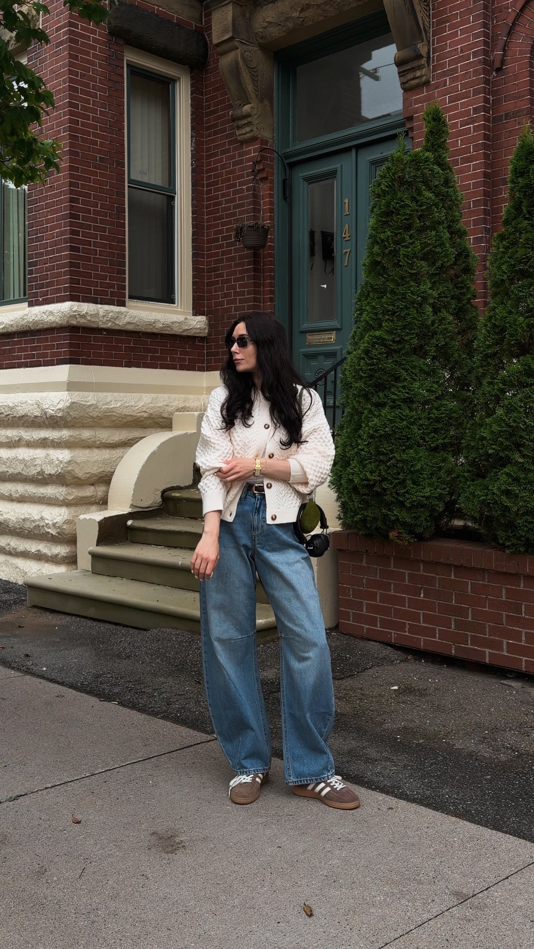 A woman stands on a city sidewalk in front of a red brick building, wearing a white cropped jacket, wide-leg blue jeans, and brown sneakers.