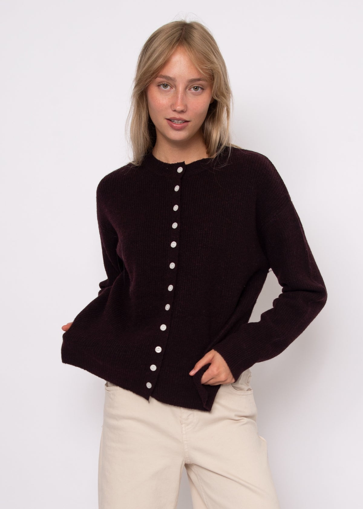 A woman modeling a dark plum button-up cardigan sweater with cream buttons, paired with beige pants, against a plain white studio background.