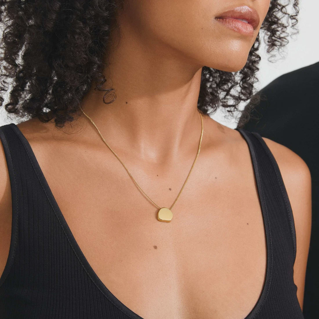 A close-up of a model wearing a delicate gold chain necklace with a small, flat, circular gold pendant. The model is wearing a black ribbed tank top, and her curly dark hair is visible against a light background.