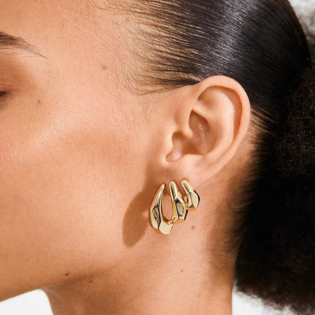 A close-up of a model's ear wearing a pair of gold crescent-shaped hoop earrings.
