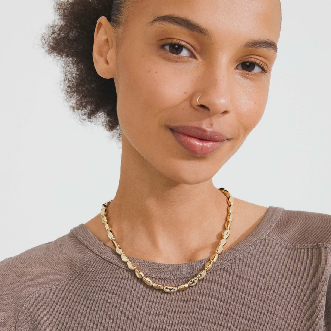 A close-up of a woman wearing a chunky gold chain necklace with textured, organic-shaped links, paired with a taupe crewneck sweatshirt against a clean white background.