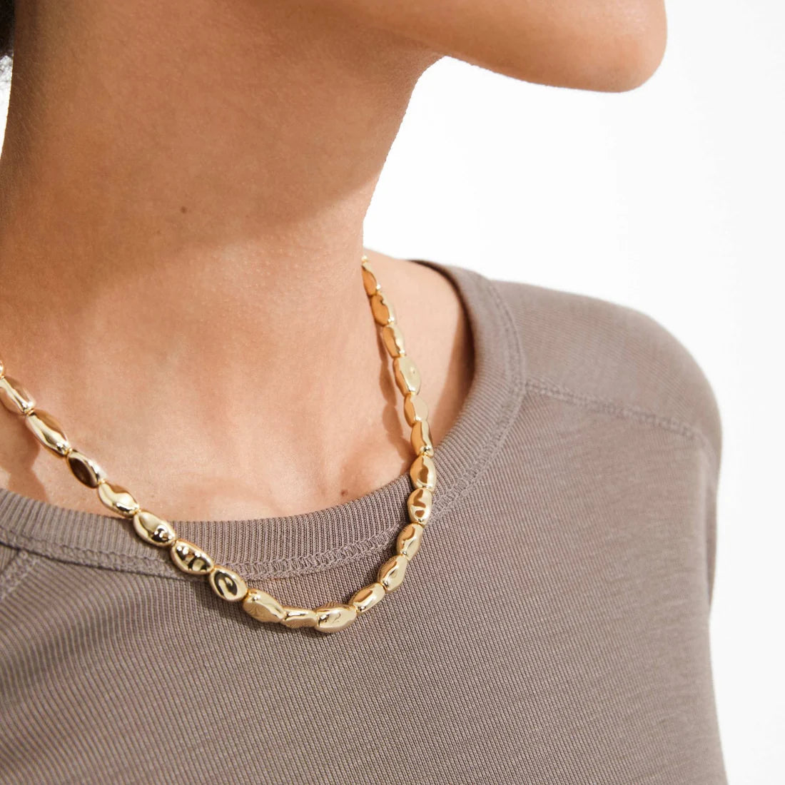 A close-up of a model wearing a polished gold chain necklace with elongated oval links over a taupe crewneck top.