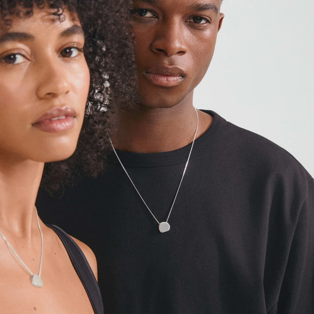 Two models wearing minimalist silver pendant necklaces with flat, geometric oval pendants against a light gray background.