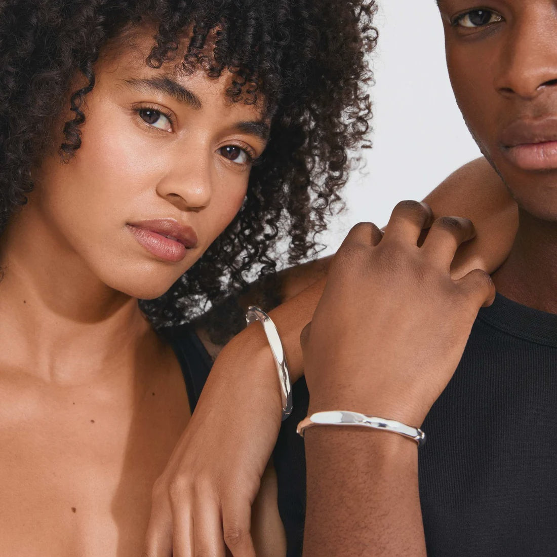 Two models wearing slim silver metal bracelets, shown in a close-up lifestyle shot against a light gray background.