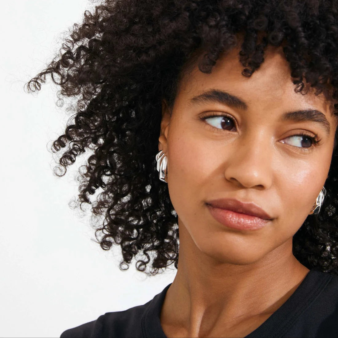 A close-up portrait of a woman with dark curly hair wearing silver hoop earrings, shown against a plain light background.