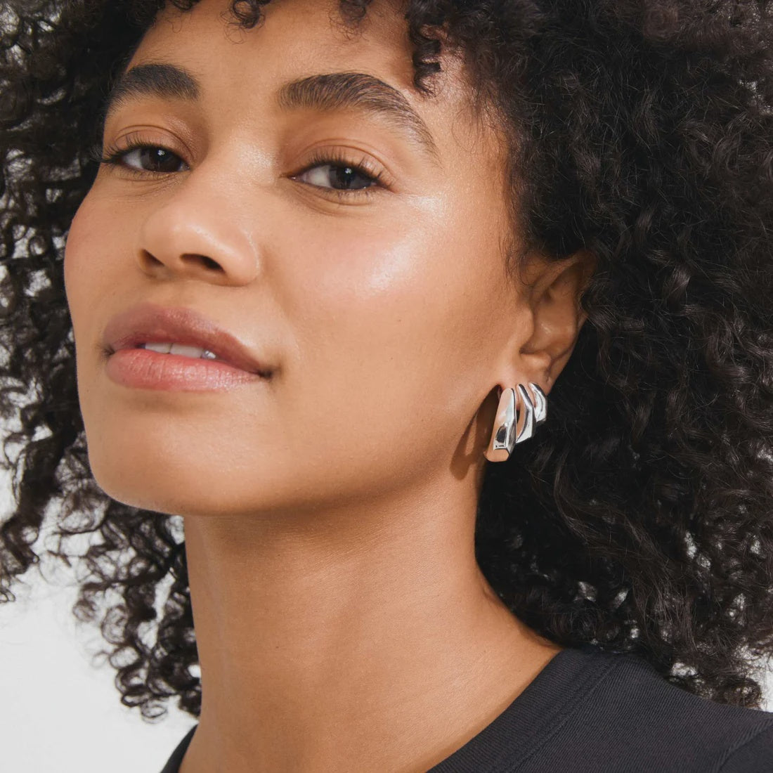 A close-up of a woman with curly dark hair wearing a silver geometric drop earring with clear stones, set against a clean white background.
