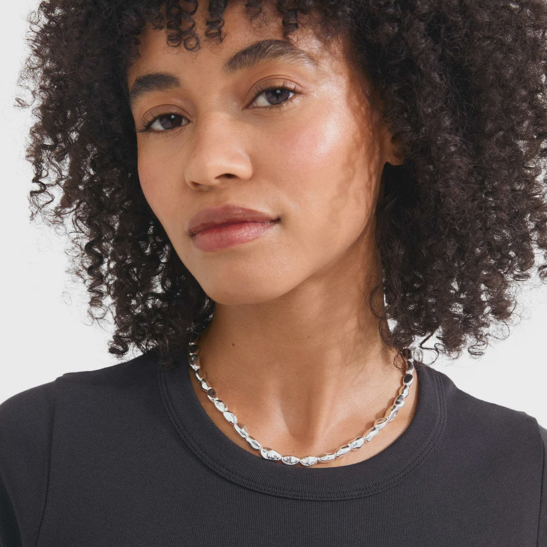 A close-up of a woman with curly dark hair wearing a silver chain necklace and a black crew-neck top against a light gray background.
