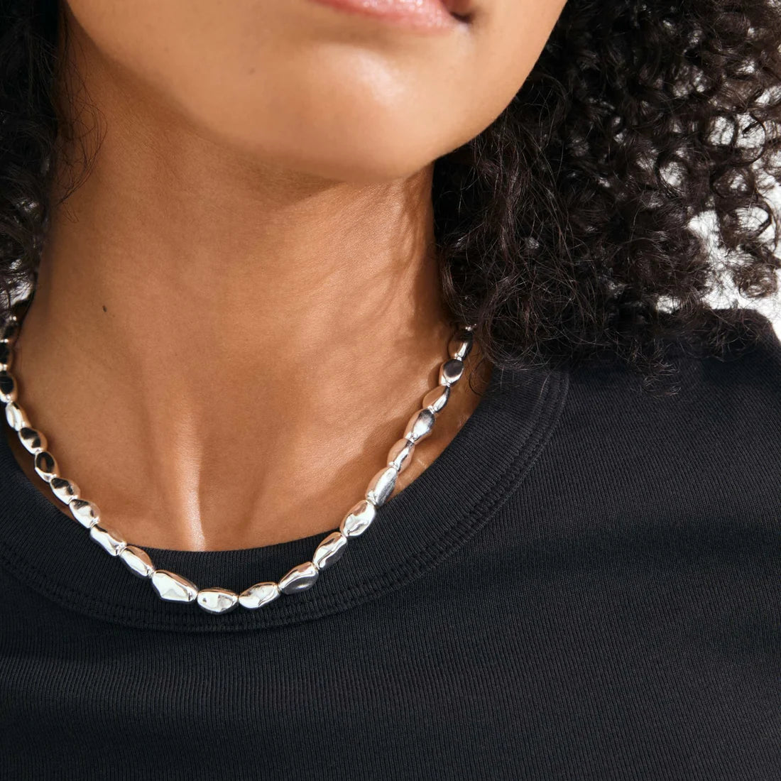 A close-up of a model wearing a chunky silver chain necklace with irregular, polished links over a black crewneck shirt.