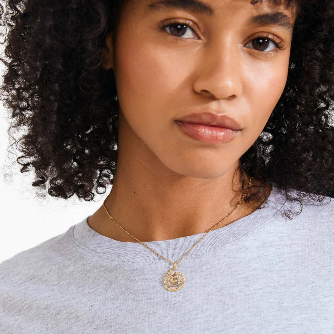 A close-up of a woman with curly dark hair wearing a delicate gold necklace with a rose-shaped pendant, paired with a light gray heathered t-shirt against a white background.