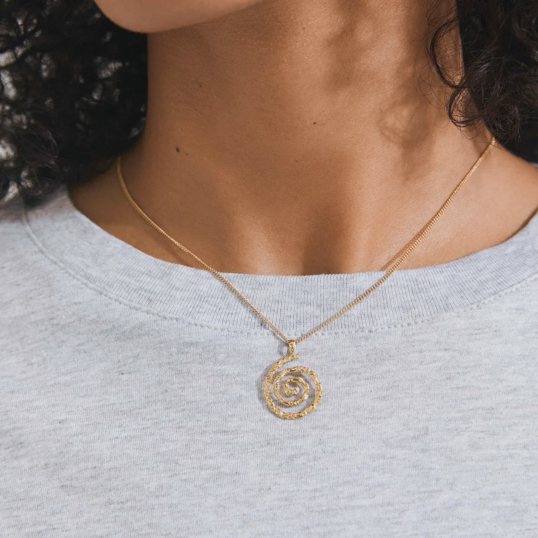 A gold spiral pendant necklace worn by a model in a light gray heathered t-shirt.