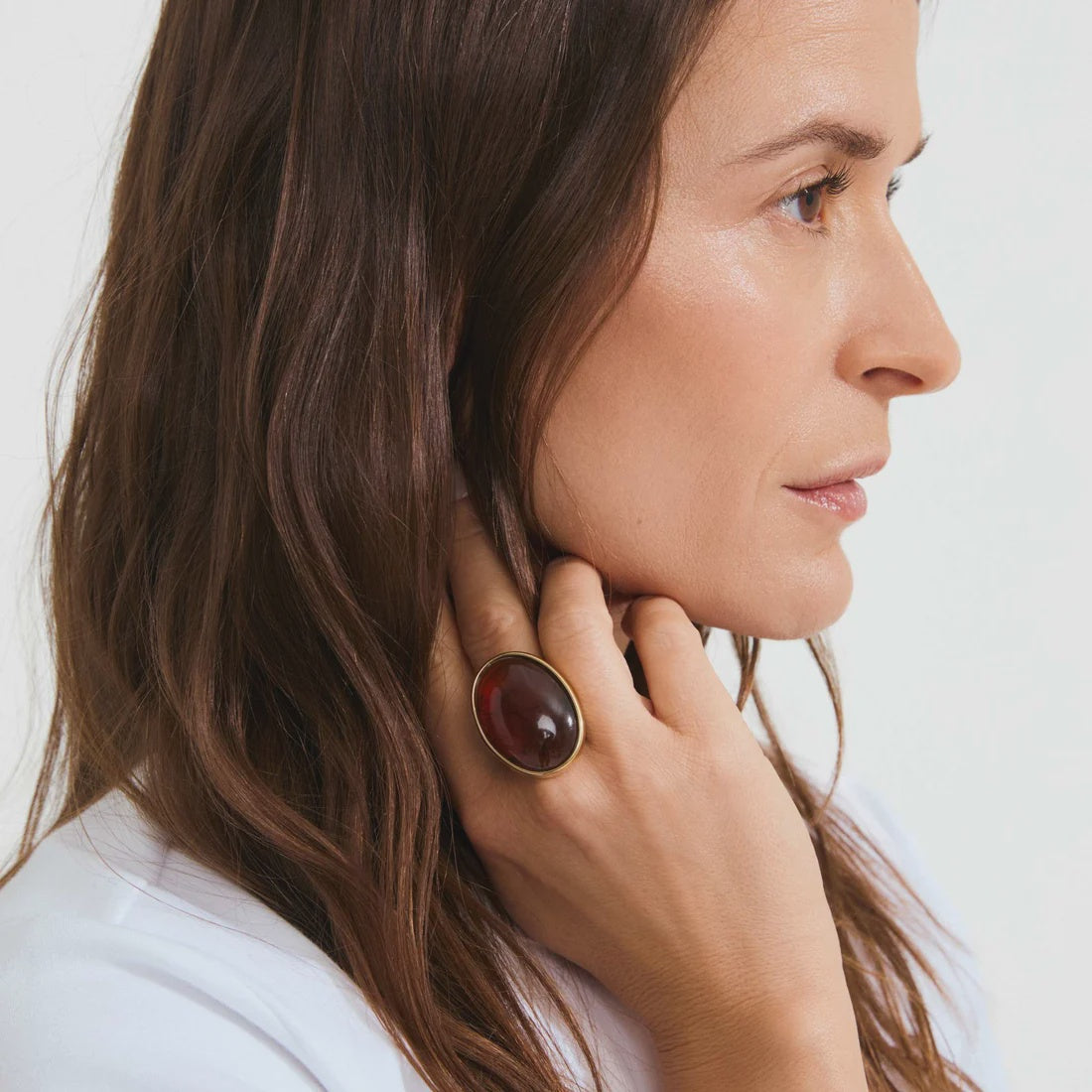A close-up side profile of a woman with long brown hair wearing a large oval ring with a dark reddish-brown stone and a gold-toned bezel on her hand.