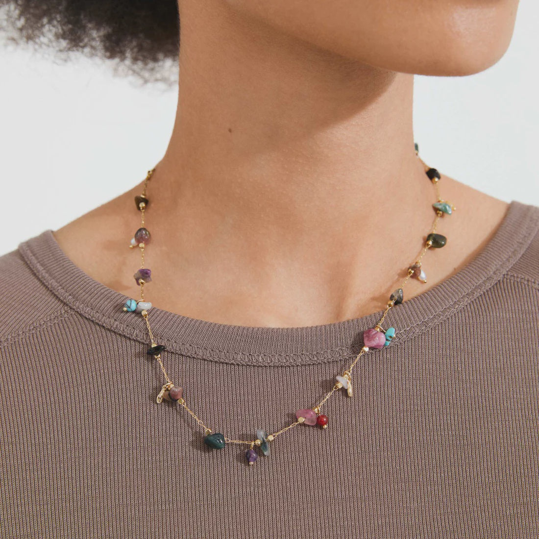 A close-up of a model wearing a delicate gold chain necklace adorned with small, colorful, irregularly shaped gemstone beads. The model is wearing a ribbed brown top against a clean, light off-white background.