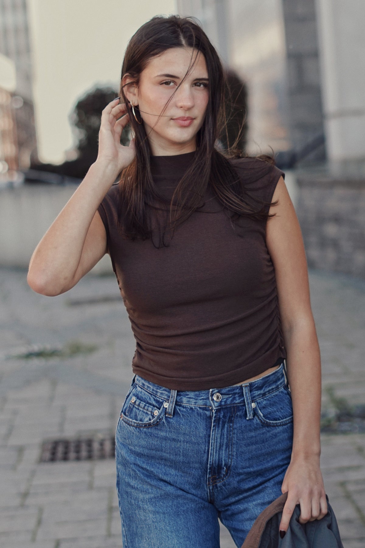 A woman wearing a fitted sleeveless brown mock-neck top and blue jeans, standing outdoors on a paved street.