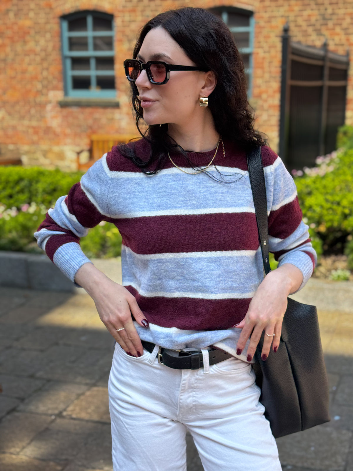 Woman wearing a burgundy and light grey striped crewneck sweater with white jeans, sunglasses and a black tote bag.