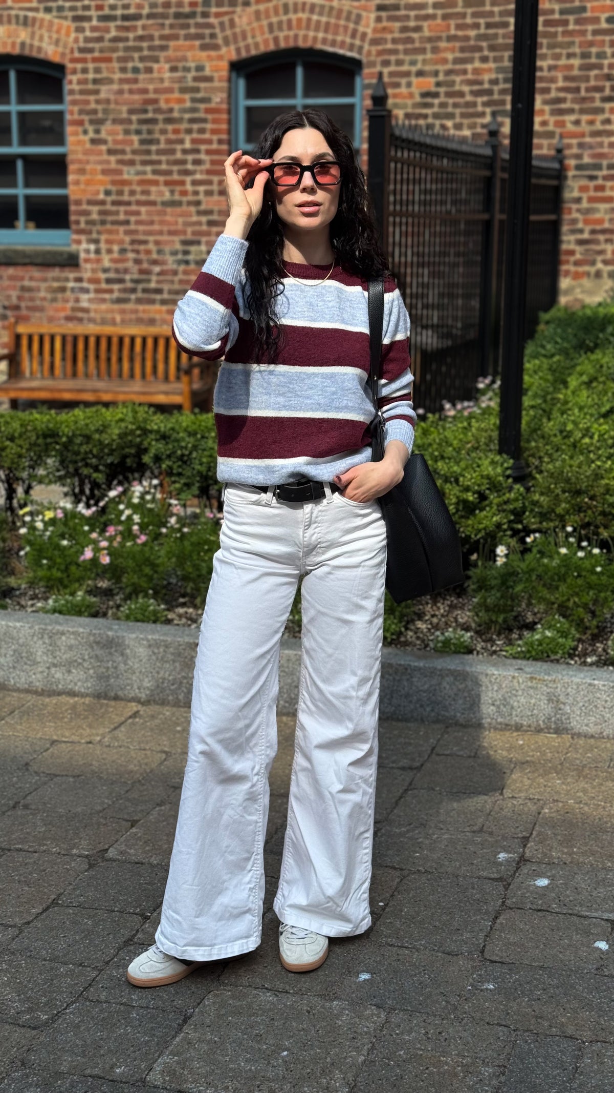 Woman wearing a burgundy and light grey striped crewneck sweater with white jeans, sunglasses and a black tote bag.