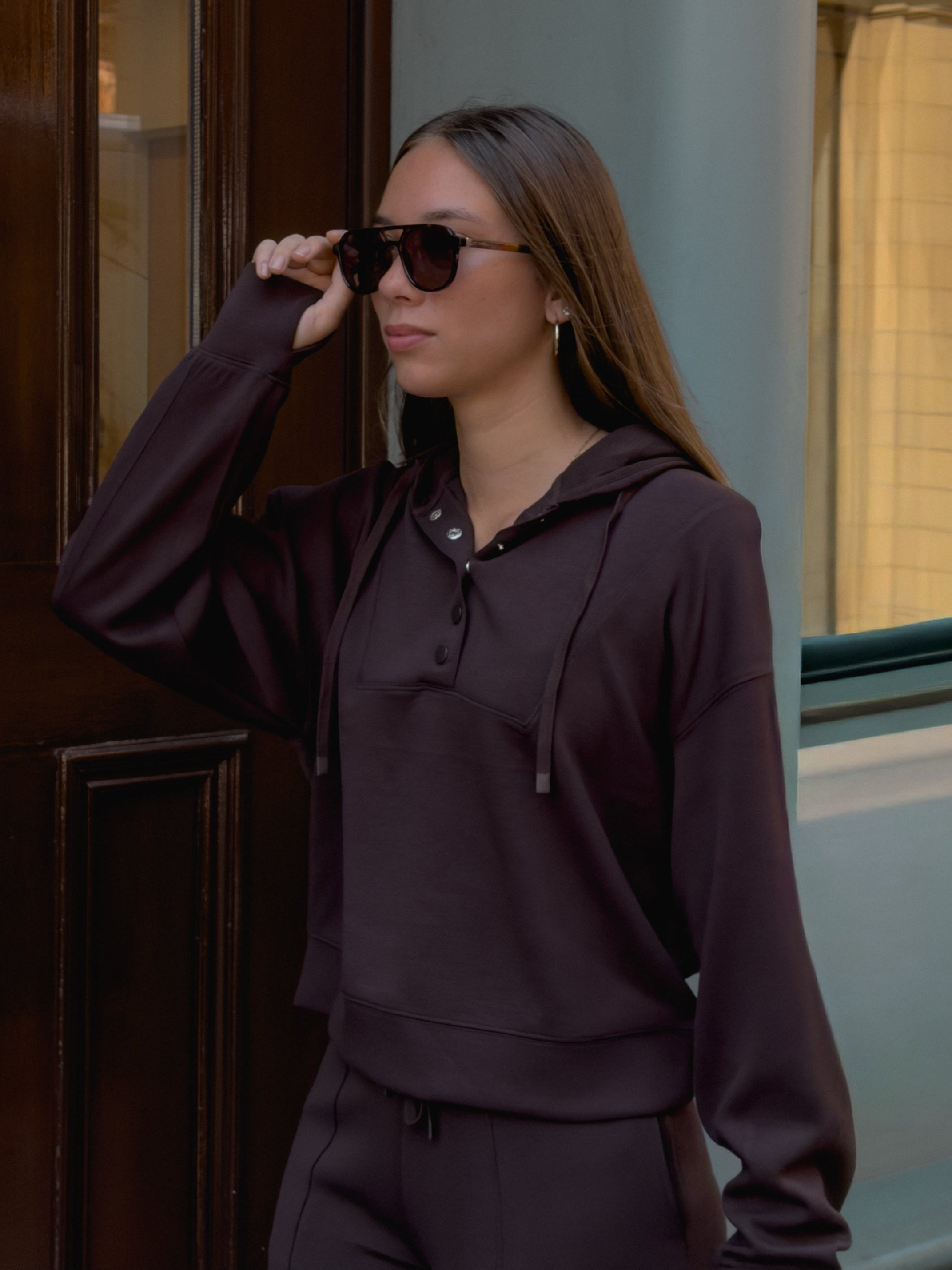 A woman wearing a dark brown matching lounge set consisting of a cropped pullover hoodie and sweatpants.