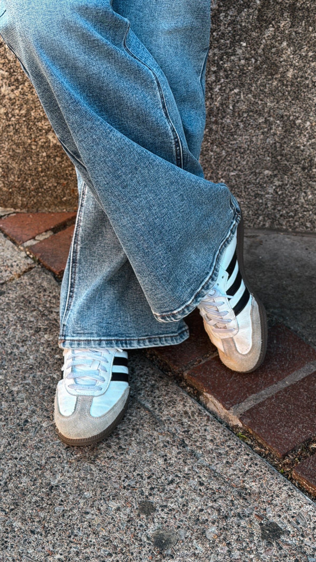 A person wearing light blue flared jeans and white Adidas Samba sneakers with black stripes, standing on a brick and concrete pavement.