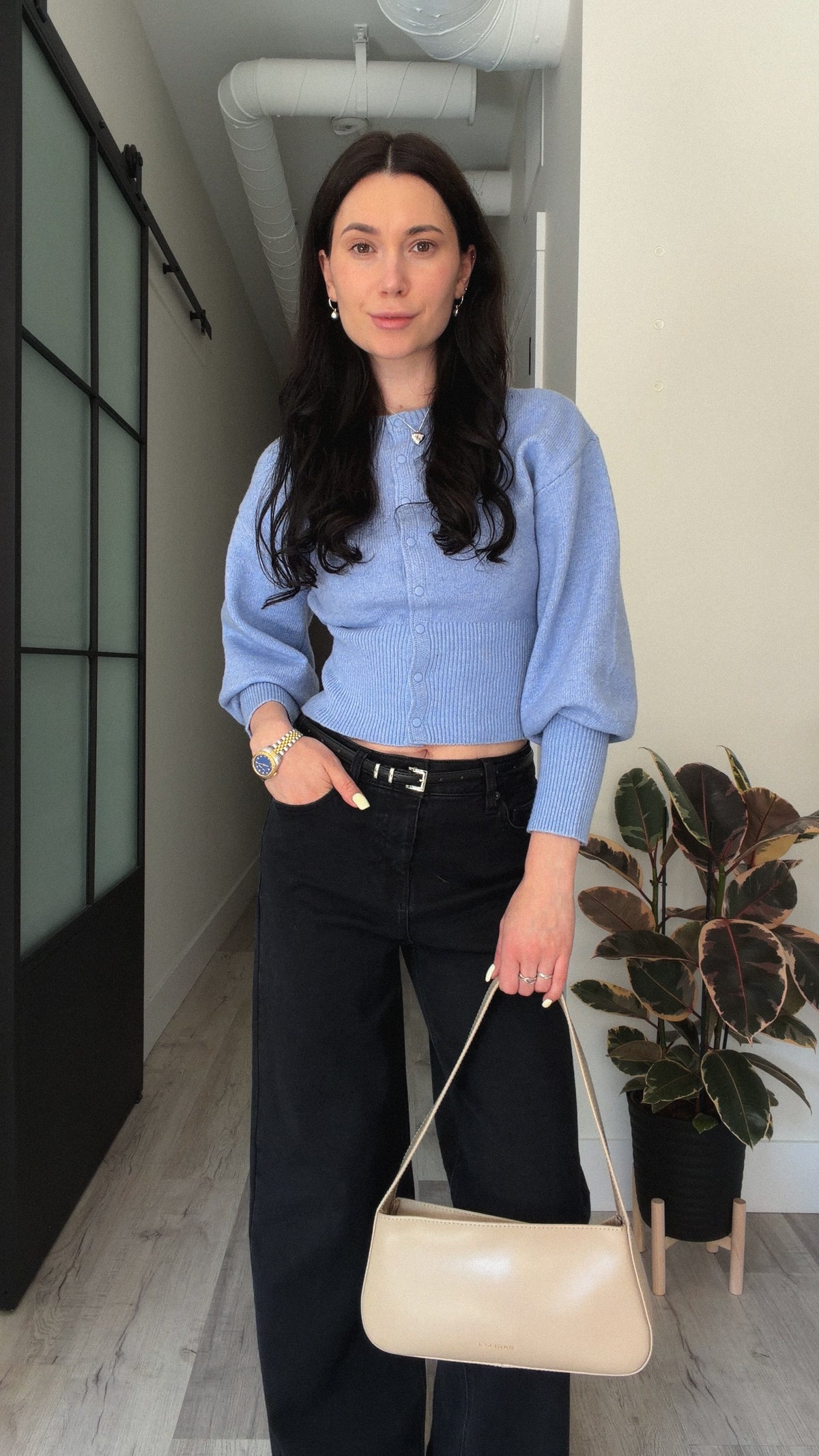A woman stands in a hallway wearing a light blue cropped knit sweater, black wide-leg pants, and holding a beige shoulder bag.