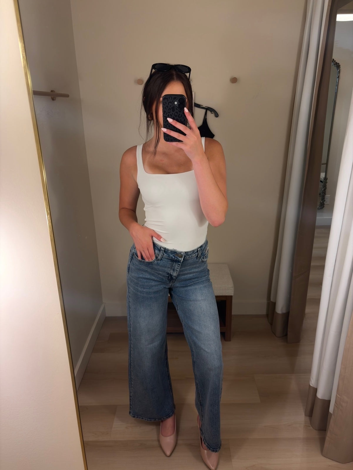A woman takes a mirror selfie in a fitting room wearing a white tank top and wide-leg blue jeans, holding a black smartphone to block the camera.
