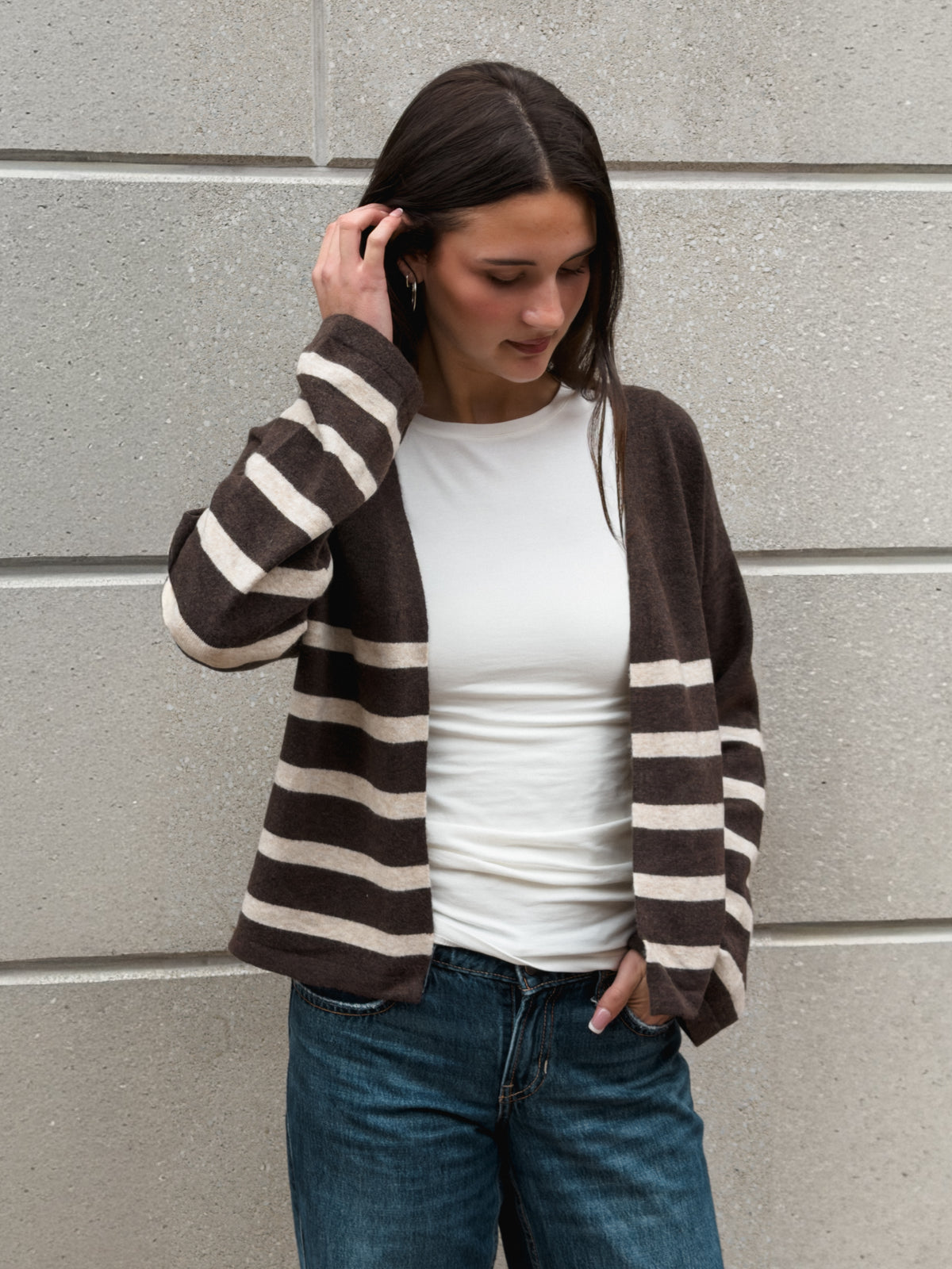 A woman wearing a brown and cream striped knit cardigan over a white t-shirt and blue jeans, standing against a gray concrete wall.