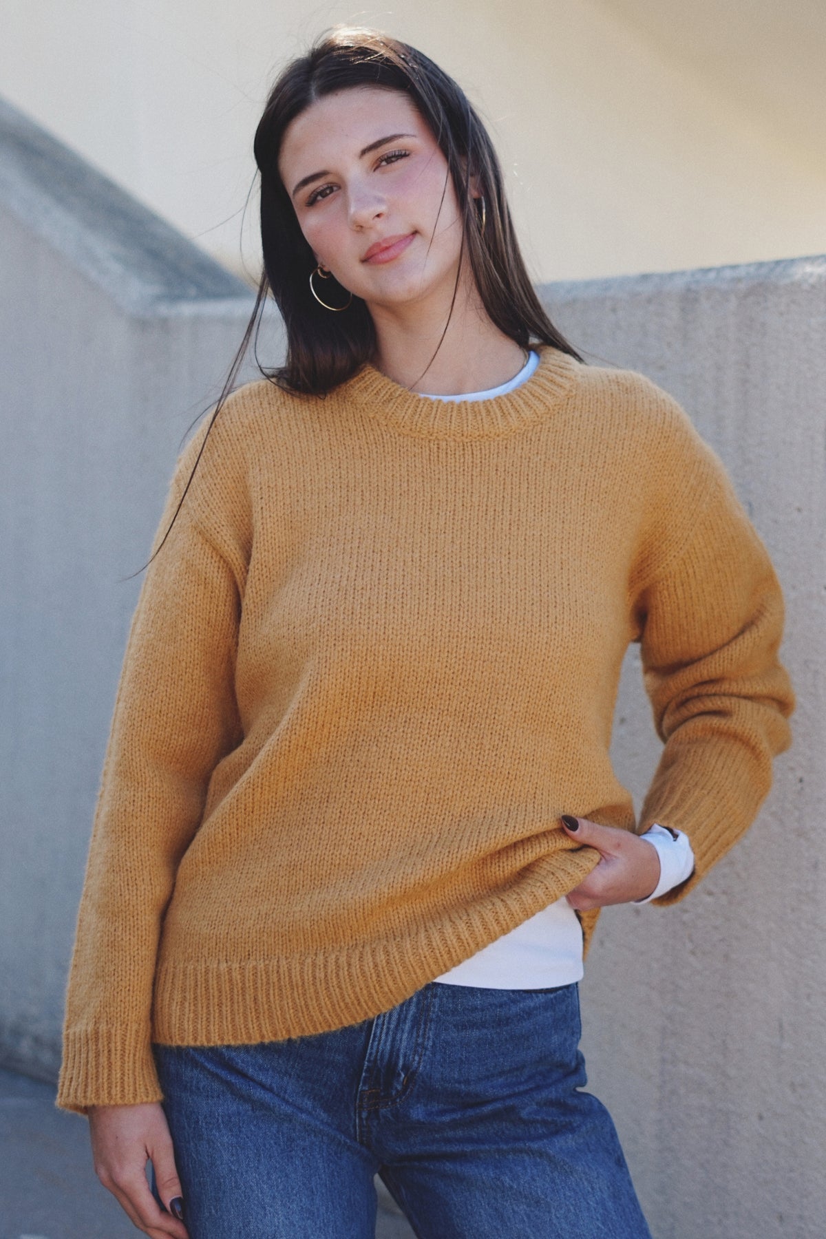 A woman wearing a mustard yellow ribbed knit crewneck sweater, blue jeans, and a white undershirt.