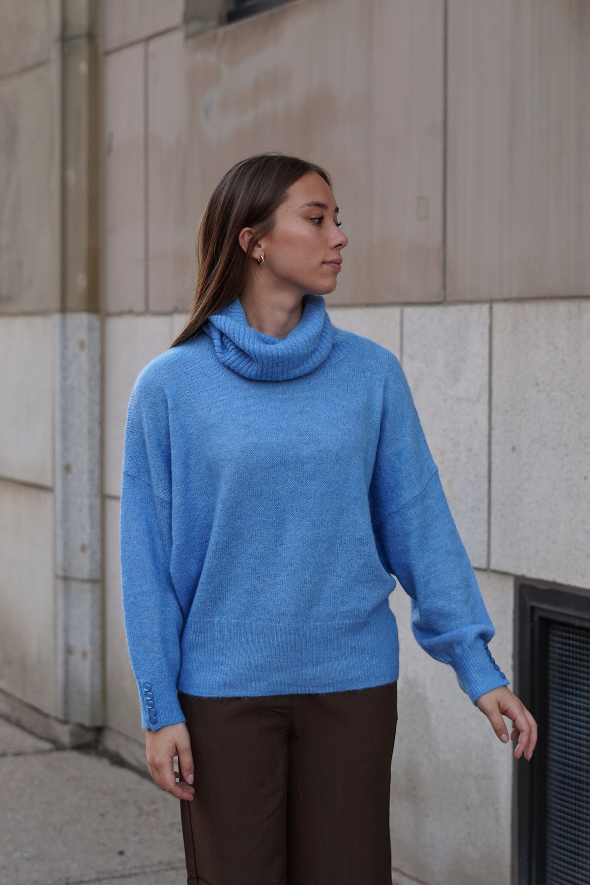 A woman wearing a light blue knit cowl-neck sweater and brown trousers, standing outdoors against a beige stone wall.