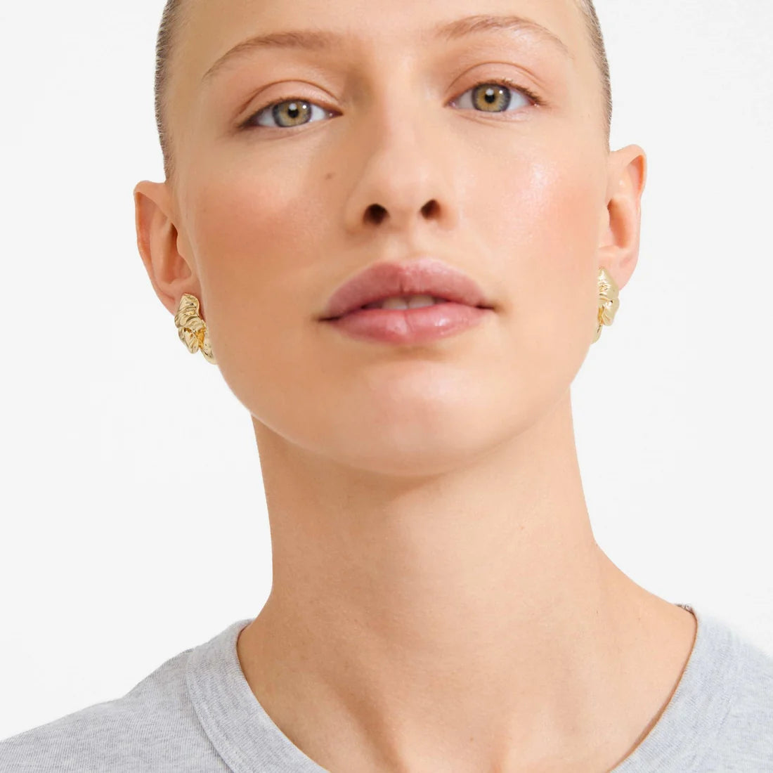 A close-up of a model wearing textured gold hoop earrings against a clean white background.